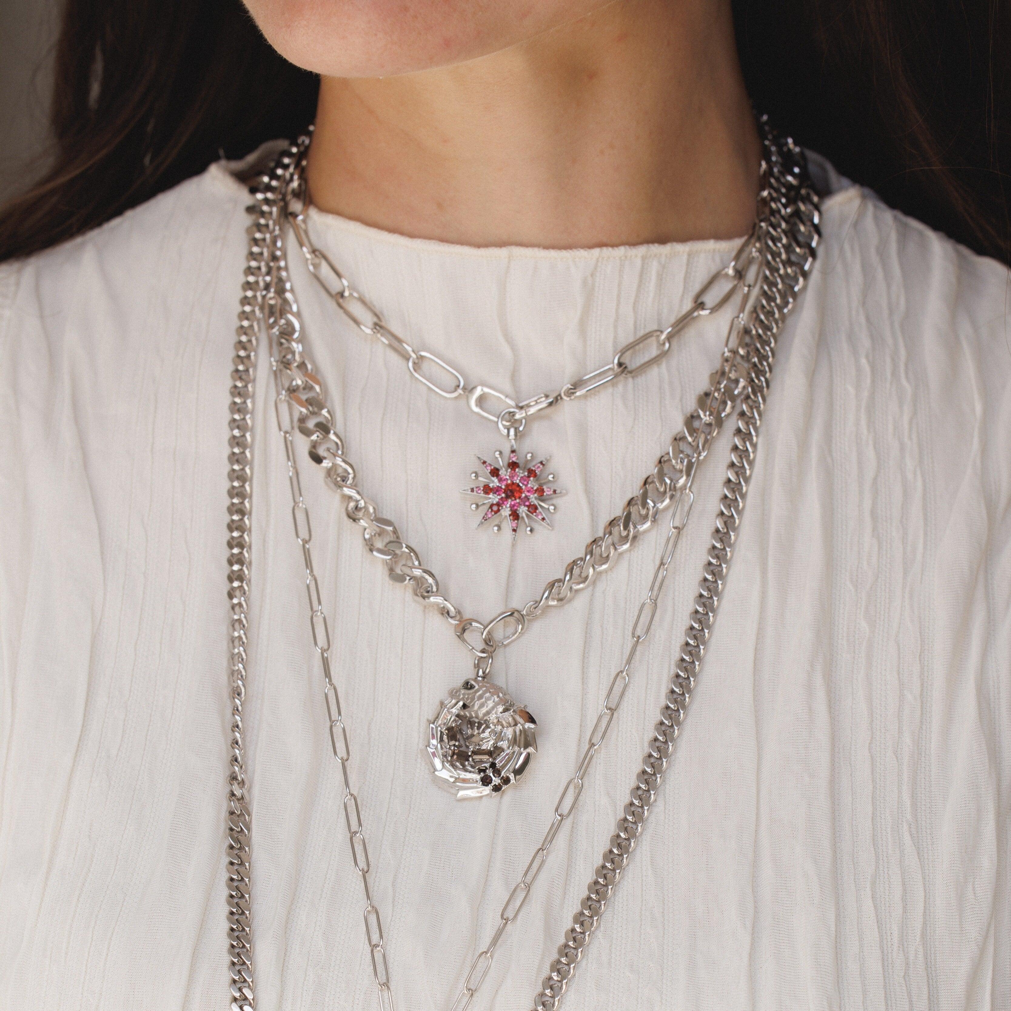 Woman in a white top wearing layered silver chain necklaces of different thicknesses and styles. She has two large silver charms hanging: a pink-and-red gemstone starburst pendant and a sculptural silver dragon-like piece.