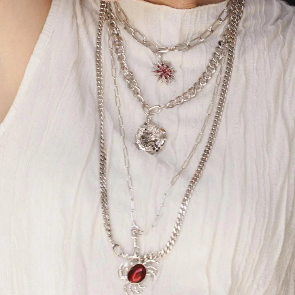 Layered silver chain necklaces featuring charms like a red starburst, dragon, and crab with red stone.