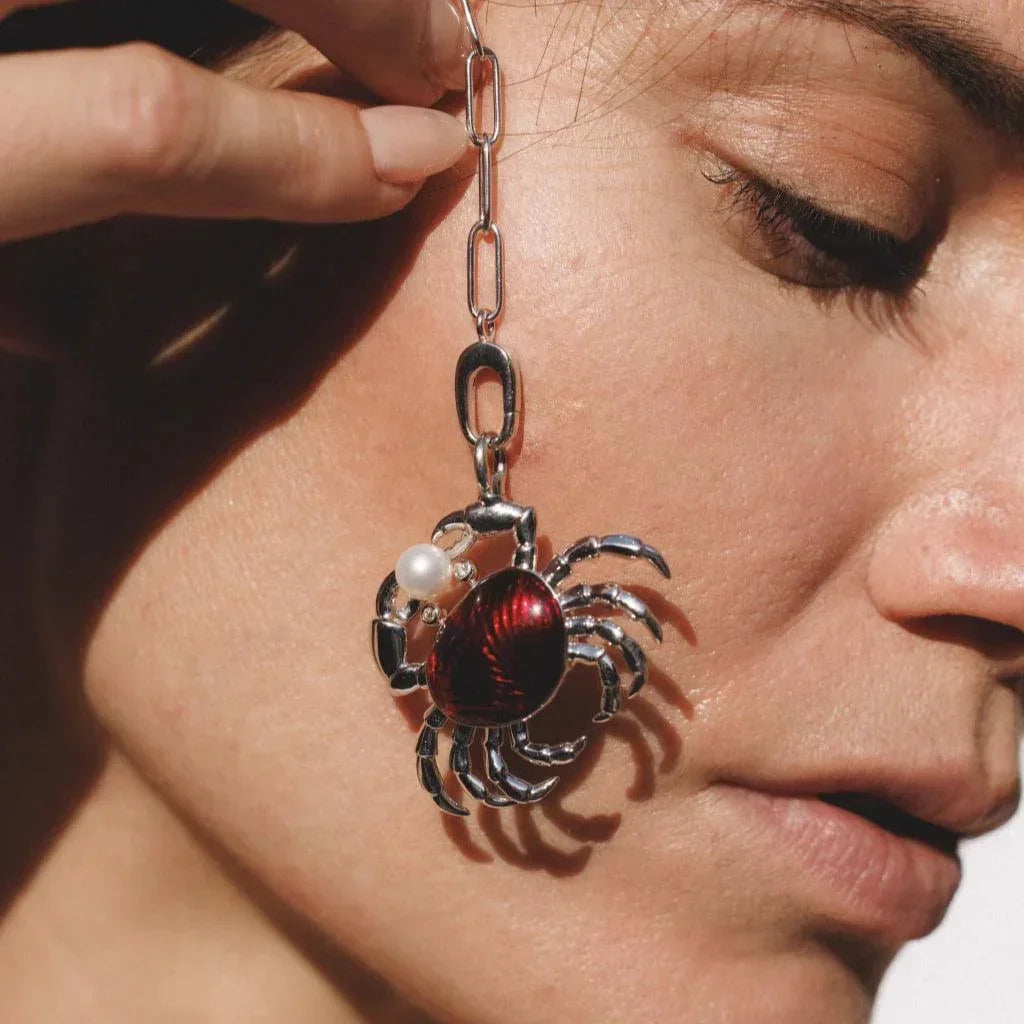 Close-up of a woman holding a silver chain adorned with a red crab charm and pearl detail.