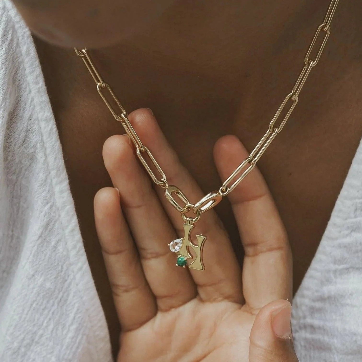 Hand holding a gold paperclip chain necklace with a gold letter H charm accented with clear and green stones.