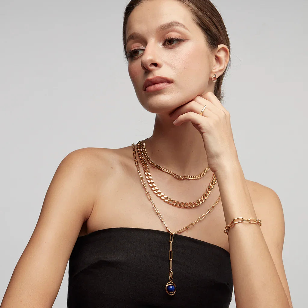 Model wearing layered gold chain necklaces with a deep blue orb pendant and matching bracelet, styled with a strapless black outfit.