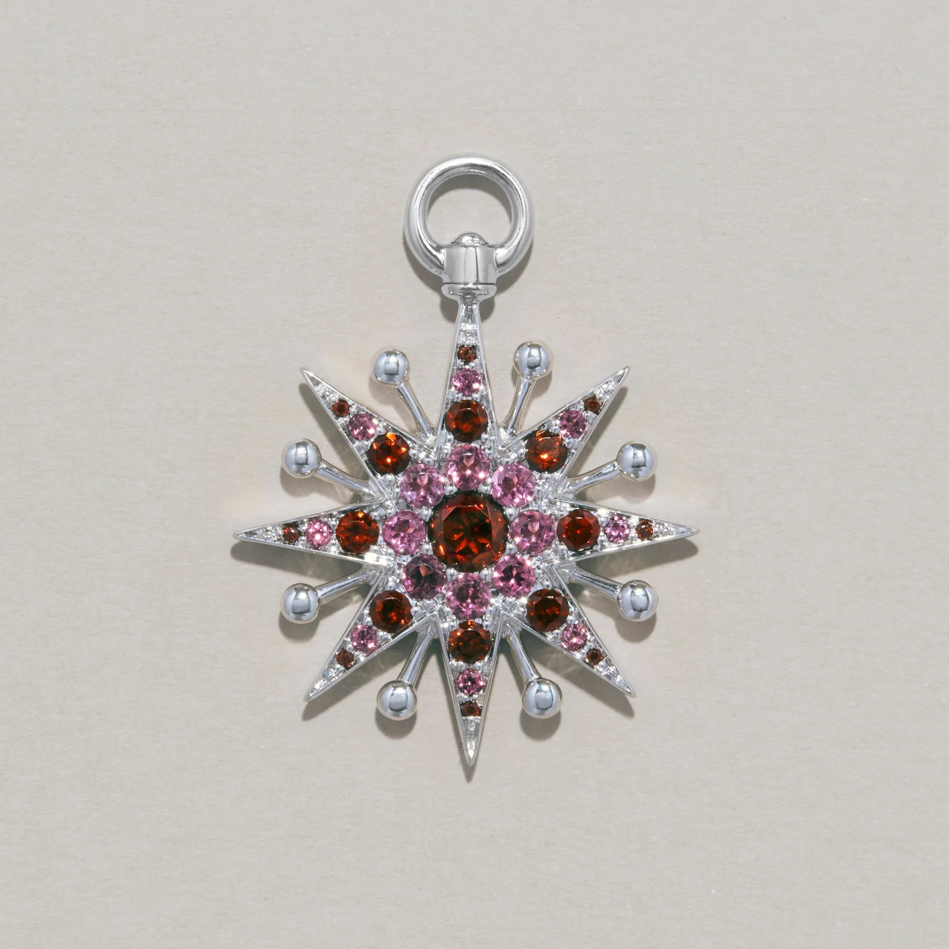 Studio product shot of a silver starburst-shaped charm set with pink and red gemstones.