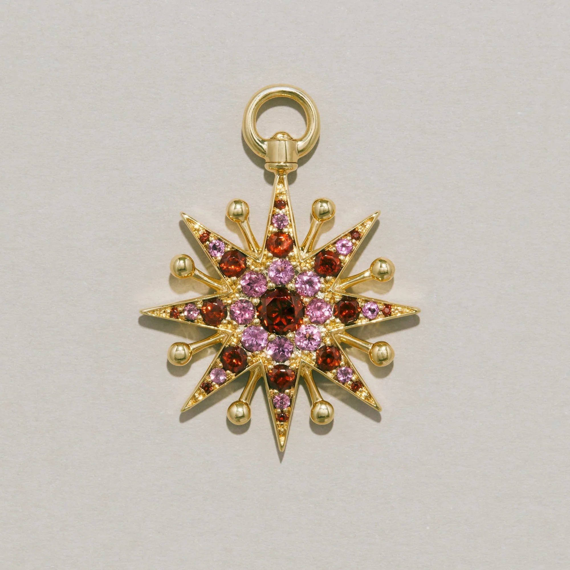 Studio product shot of a gold starburst-shaped pendant set with pink and red gemstones. The charm has radiating points tipped with gold beads and a circular bail at the top.