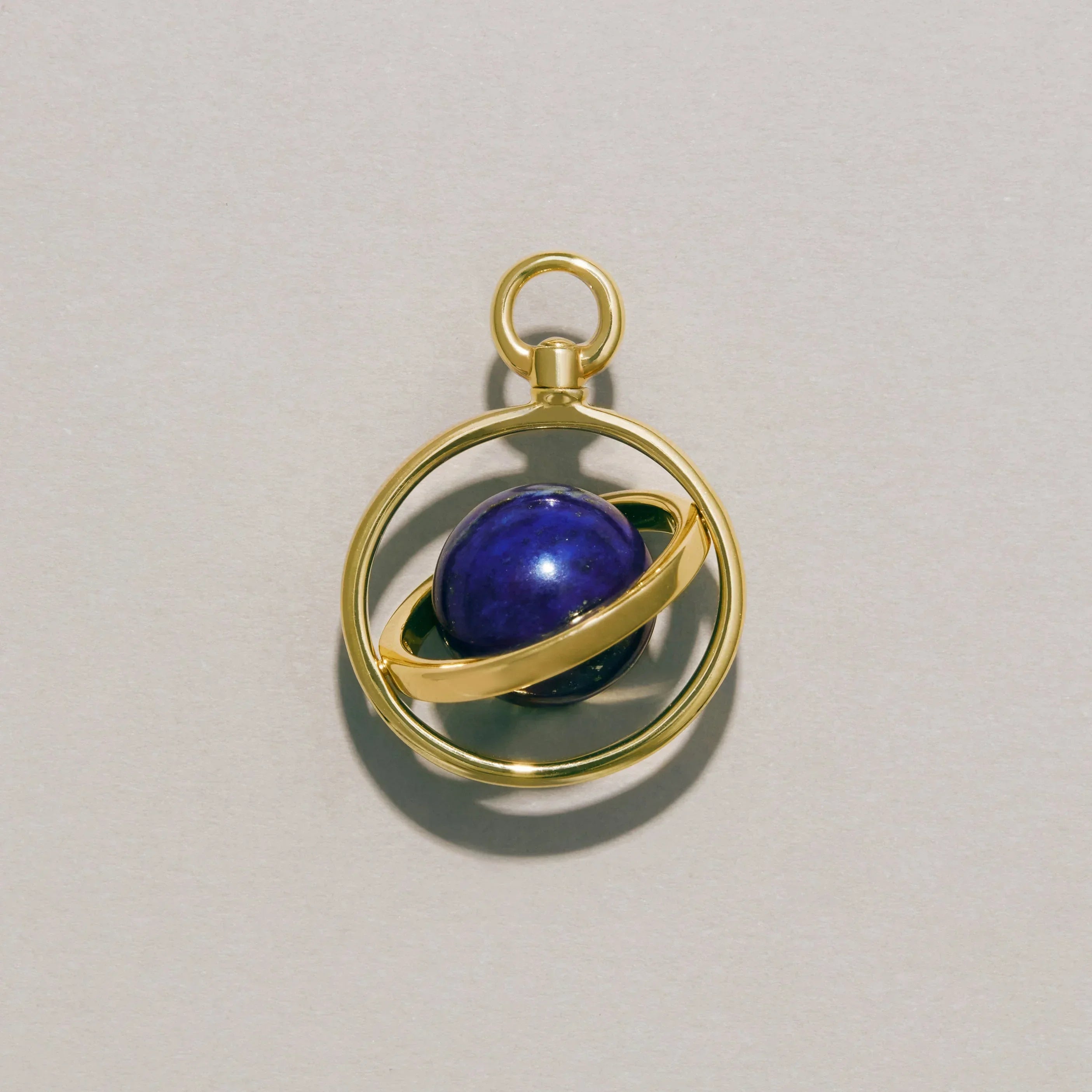 Close-up of a gold orbit-style planetary charm with a lapis lazuli sphere centerpiece resting on a neutral background.