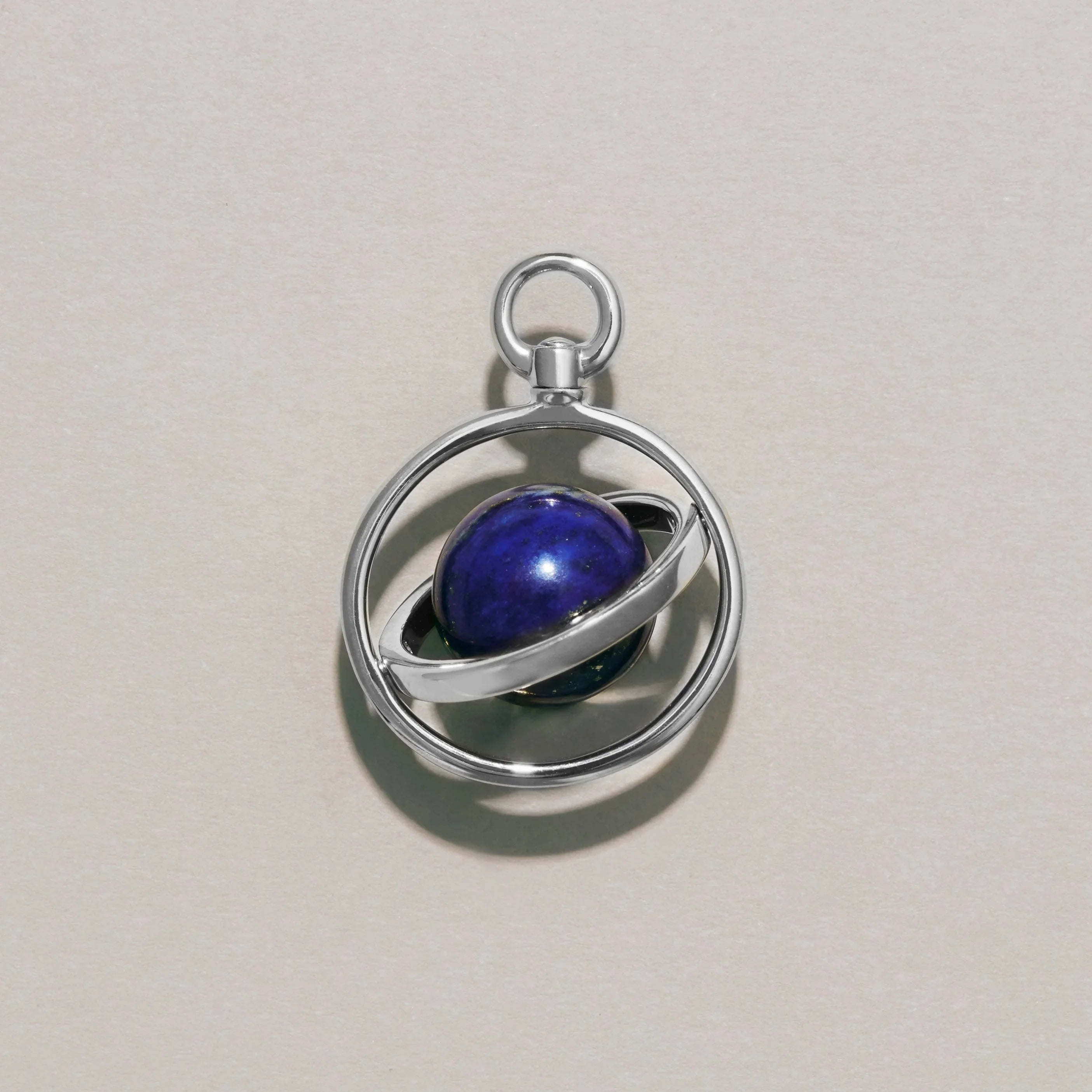 Close-up of a silver orbit-style planetary charm with a lapis lazuli sphere centerpiece resting on a neutral background.