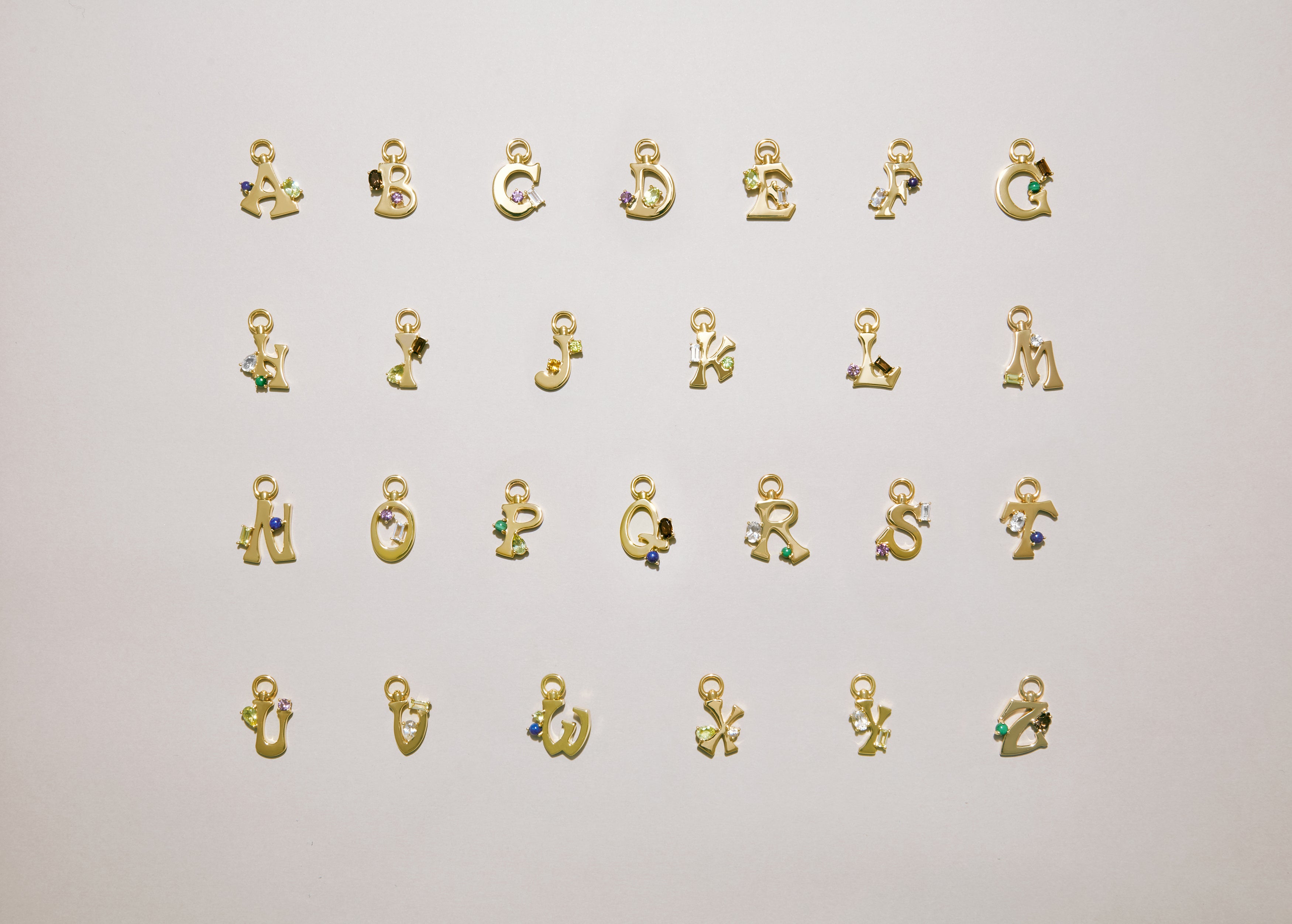 Alphabet of gold letter charms A–Z laid out on a light background, each set with colorful gemstones.