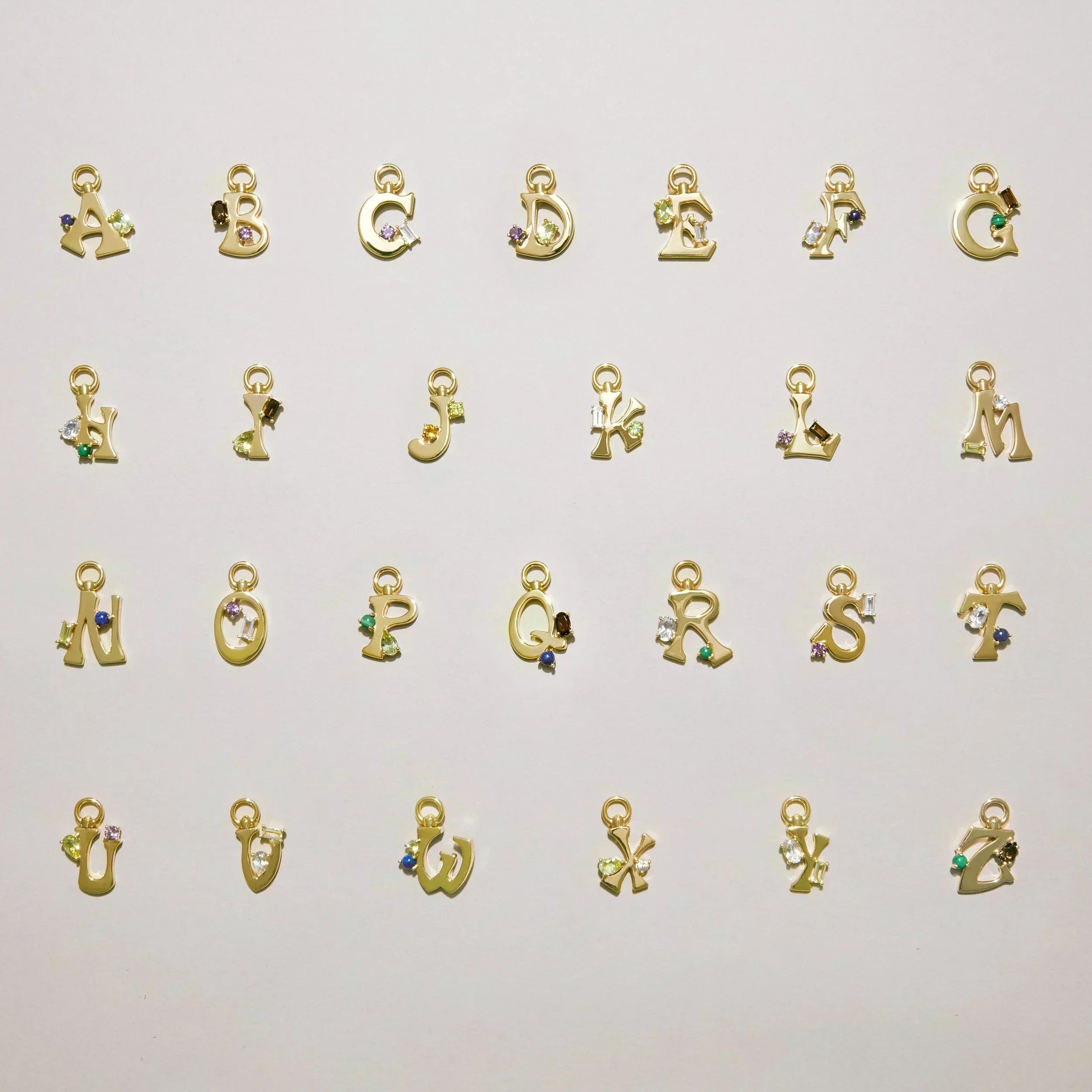 Alphabet of gold letter charms A–Z laid out on a light background, each set with colorful gemstones.