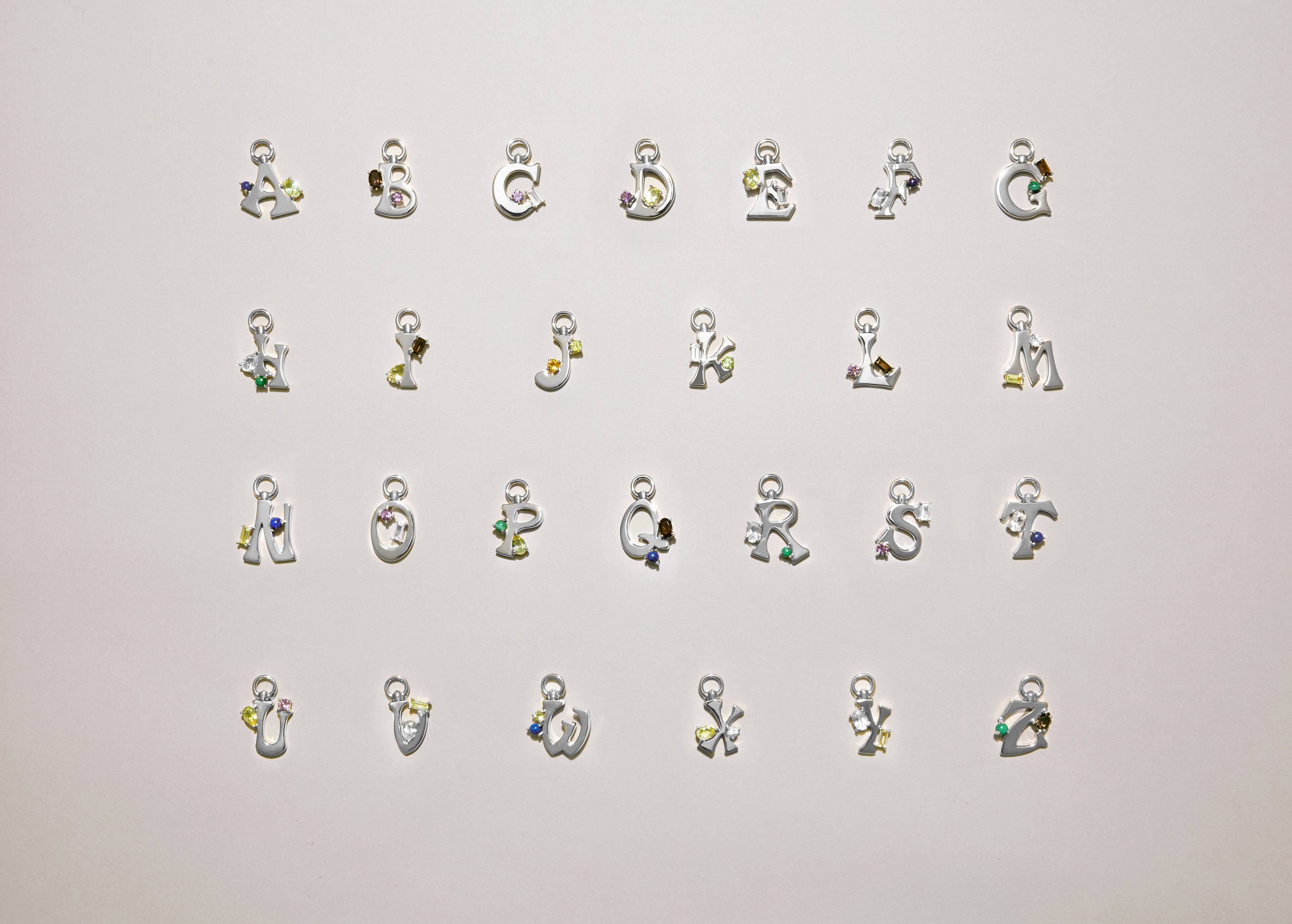 Alphabet of silver letter charms A–Z laid out on a light background, each set with colorful gemstones.