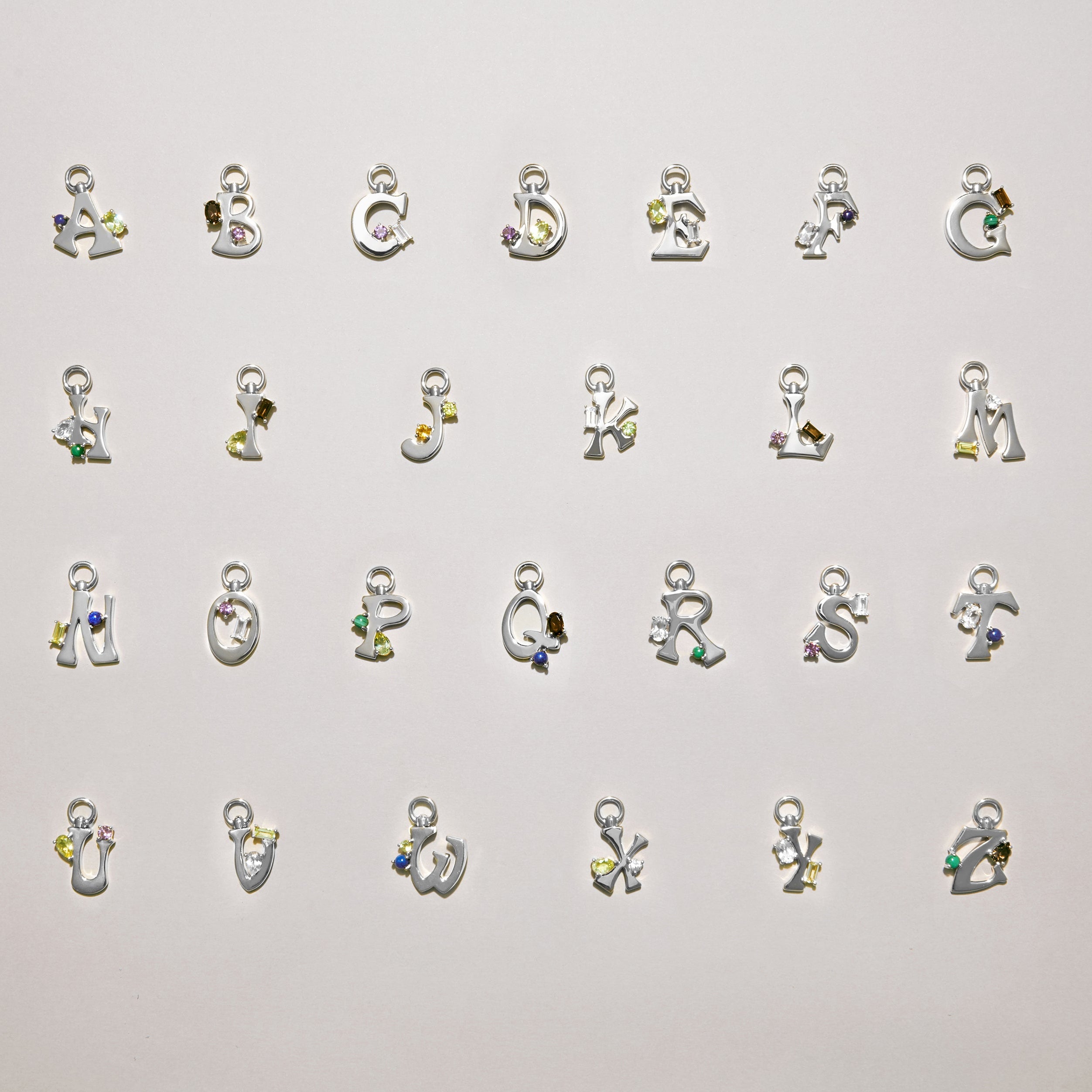 Alphabet of silver letter charms A–Z laid out on a light background, each set with colorful gemstones.