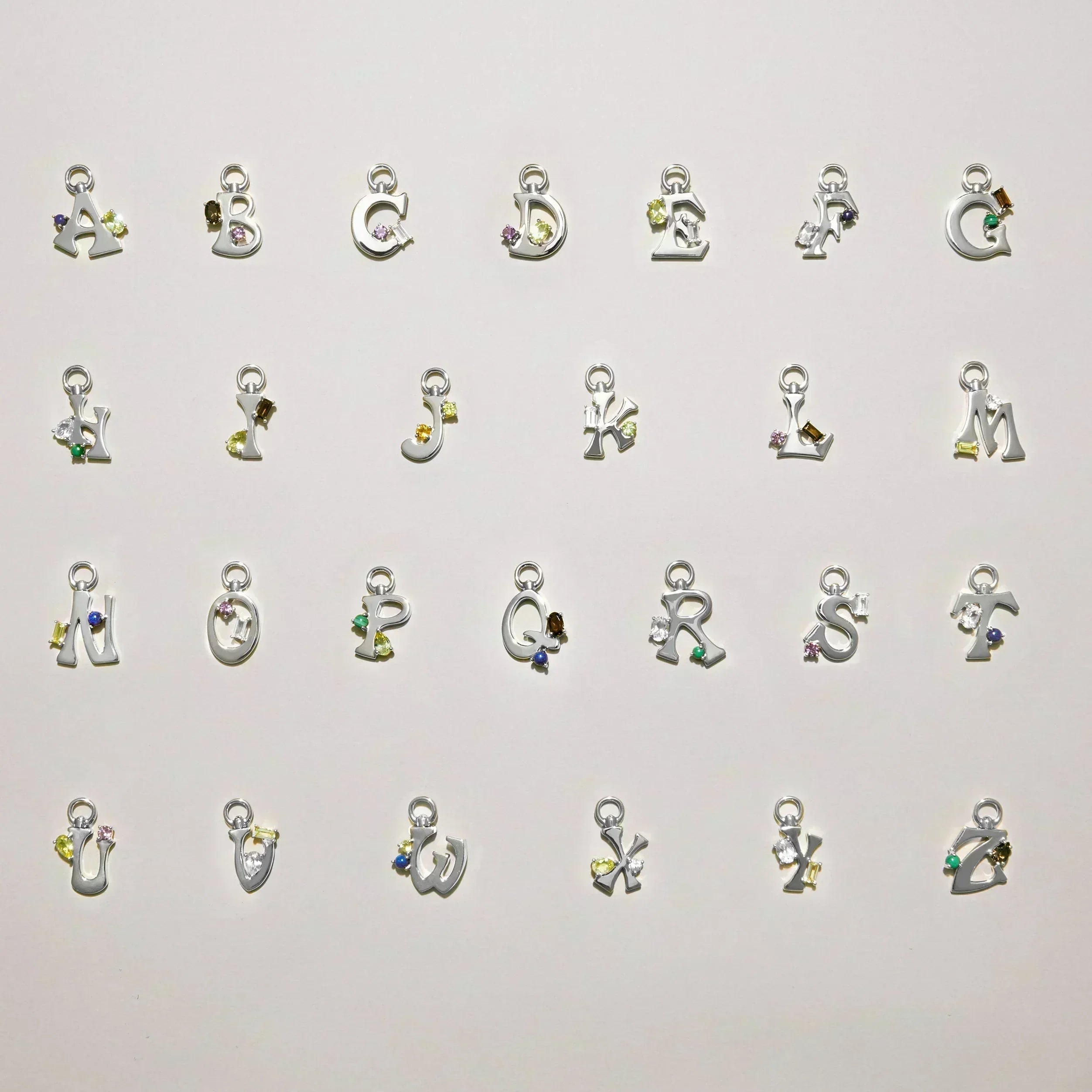 Alphabet of silver letter charms A–Z laid out on a light background, each set with colorful gemstones.