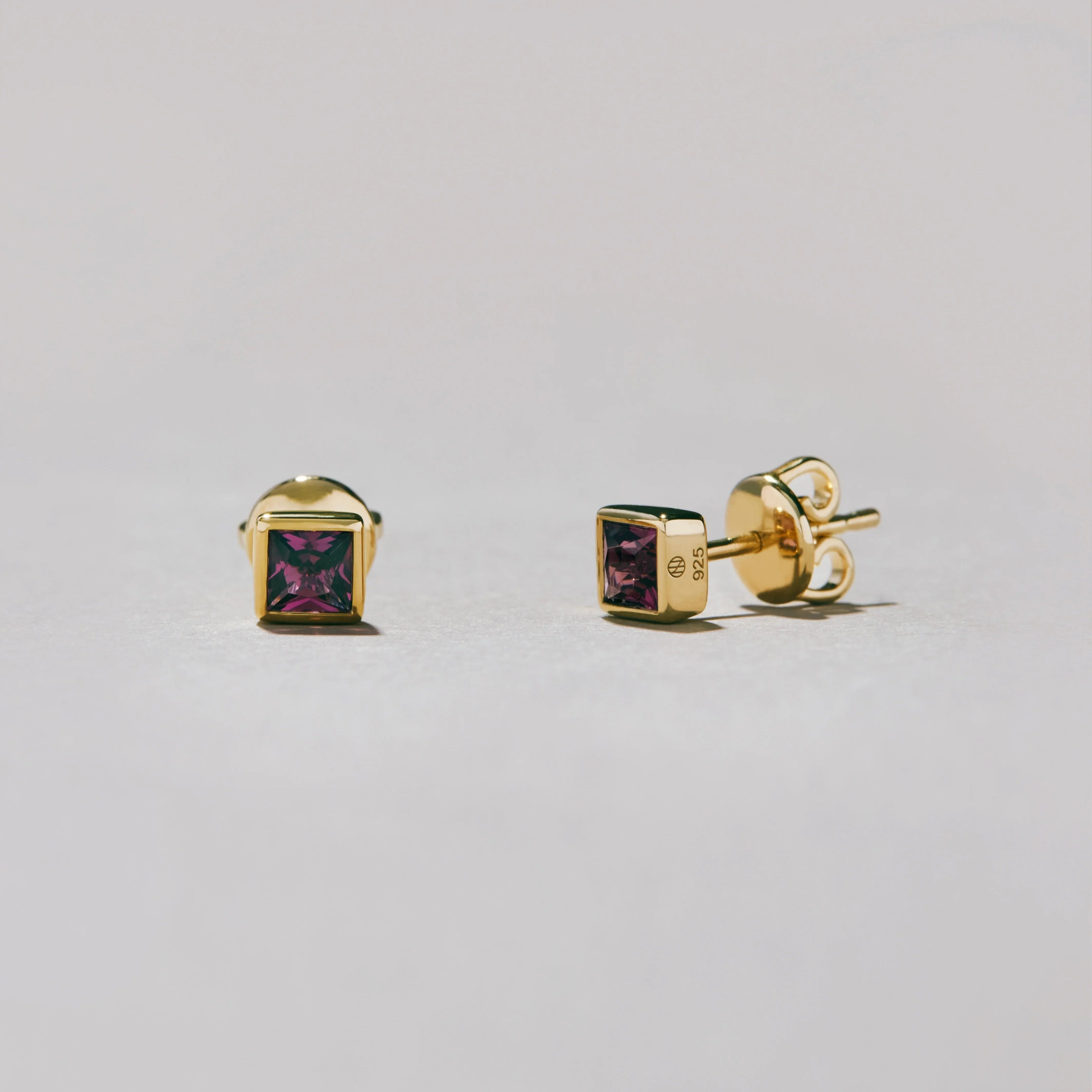 Gold square stud earrings with purple gemstones on a light gray background