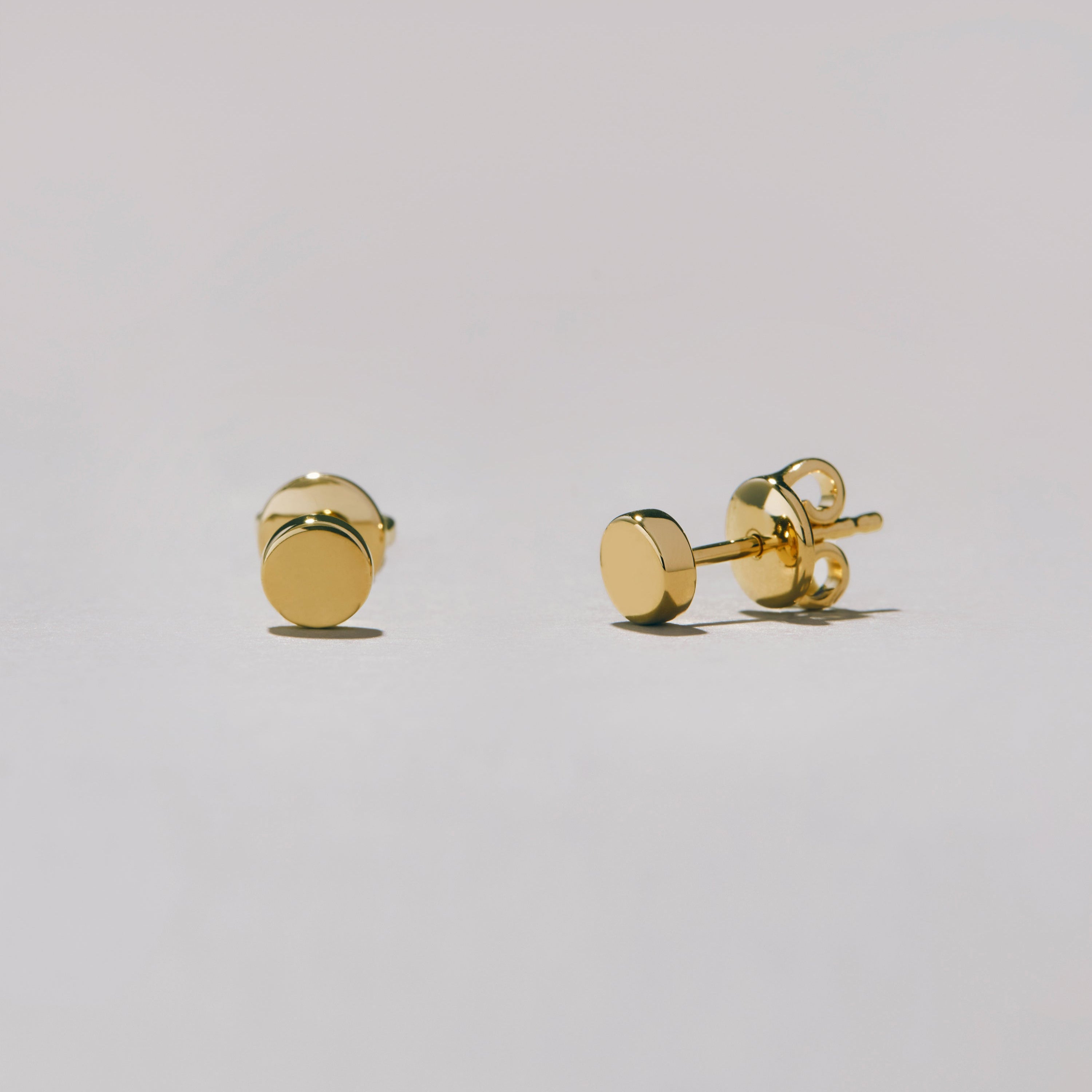 Studio shot of a pair of gold-tone round dot stud earrings lying on a white surface, shown at an angle to highlight their glossy finish.