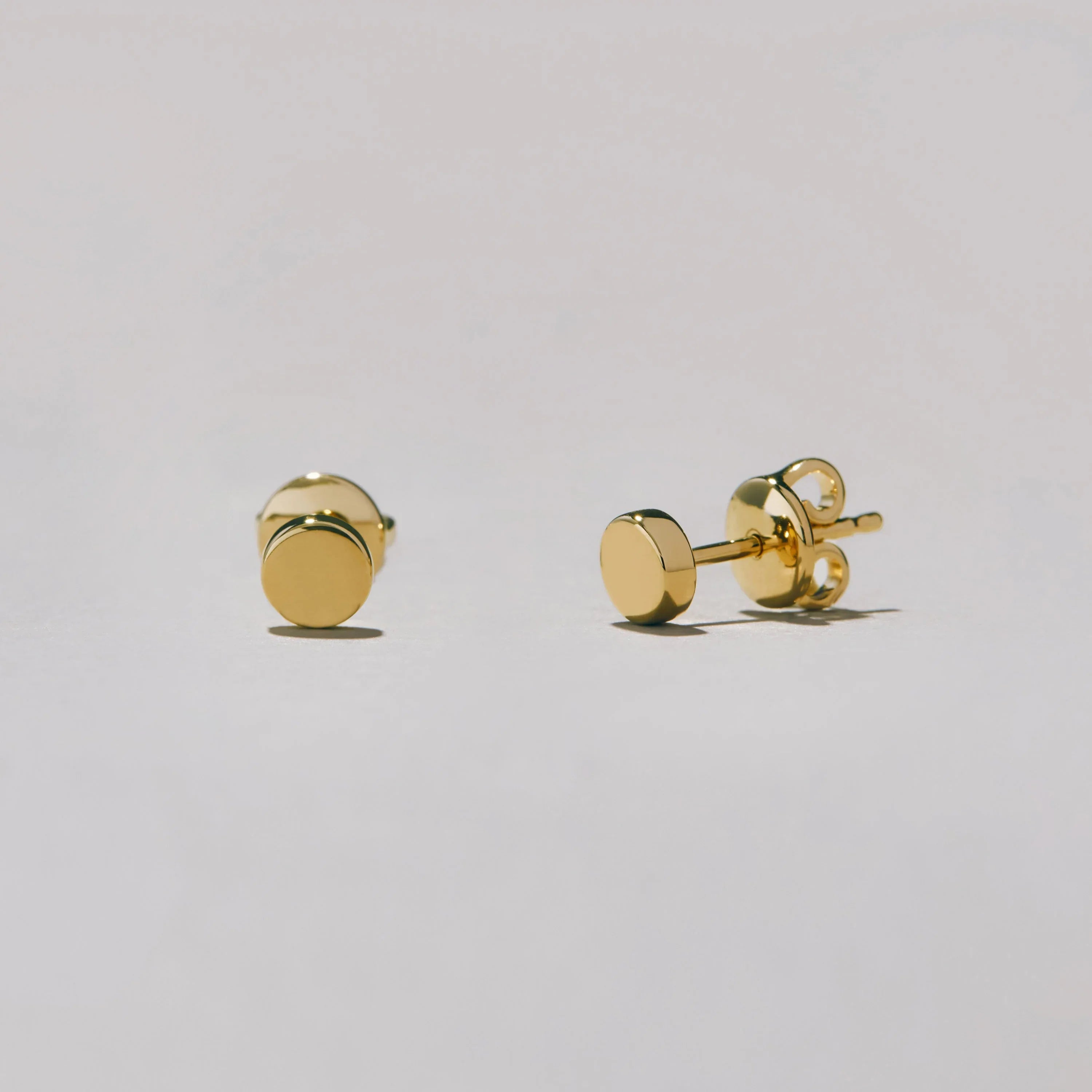 Studio shot of a pair of gold-tone round dot stud earrings lying on a white surface, shown at an angle to highlight their glossy finish.