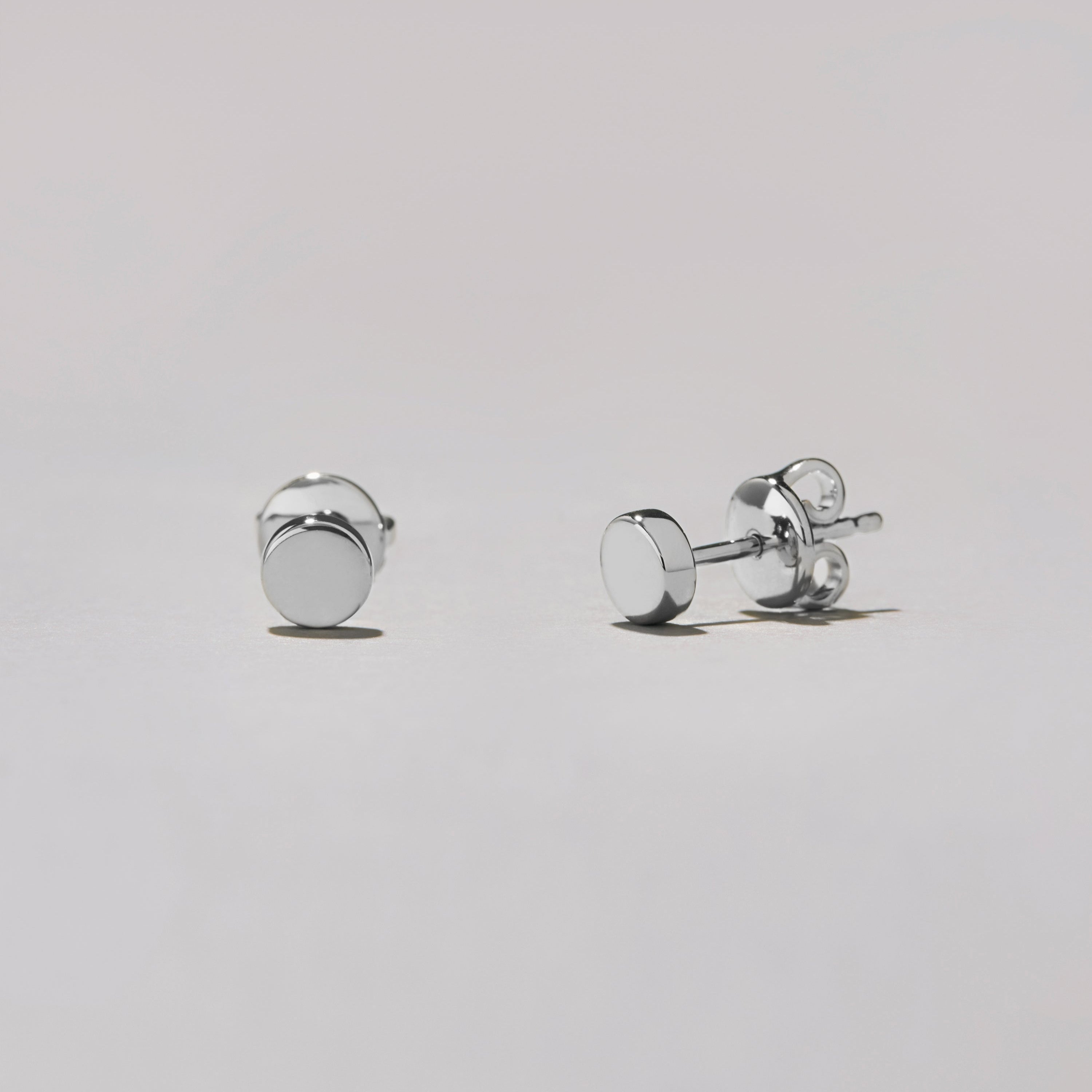 Studio shot of a pair of silver-tone round dot stud earrings lying on a white surface, shown at an angle to highlight their glossy finish.