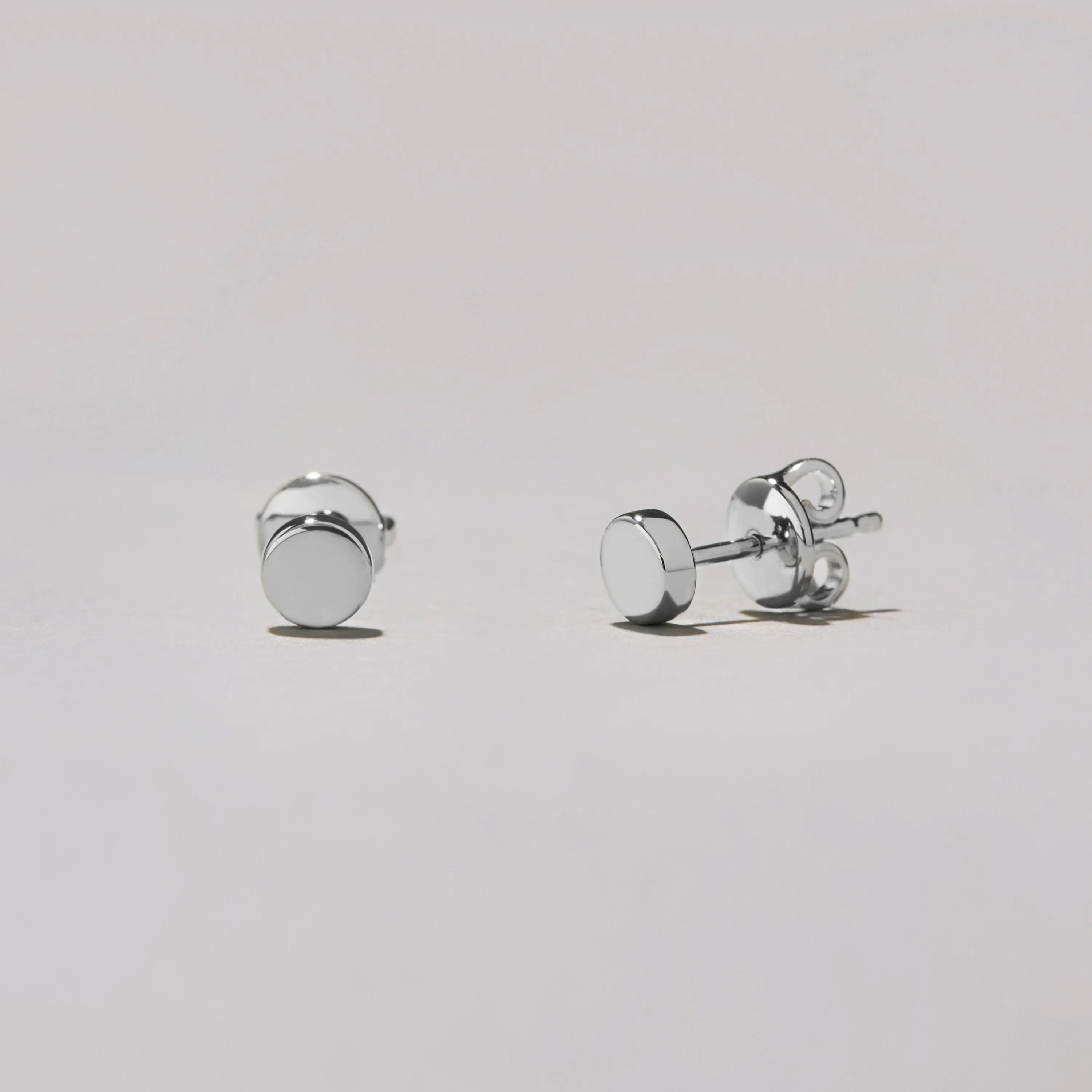 Studio shot of a pair of silver-tone round dot stud earrings lying on a white surface, shown at an angle to highlight their glossy finish.