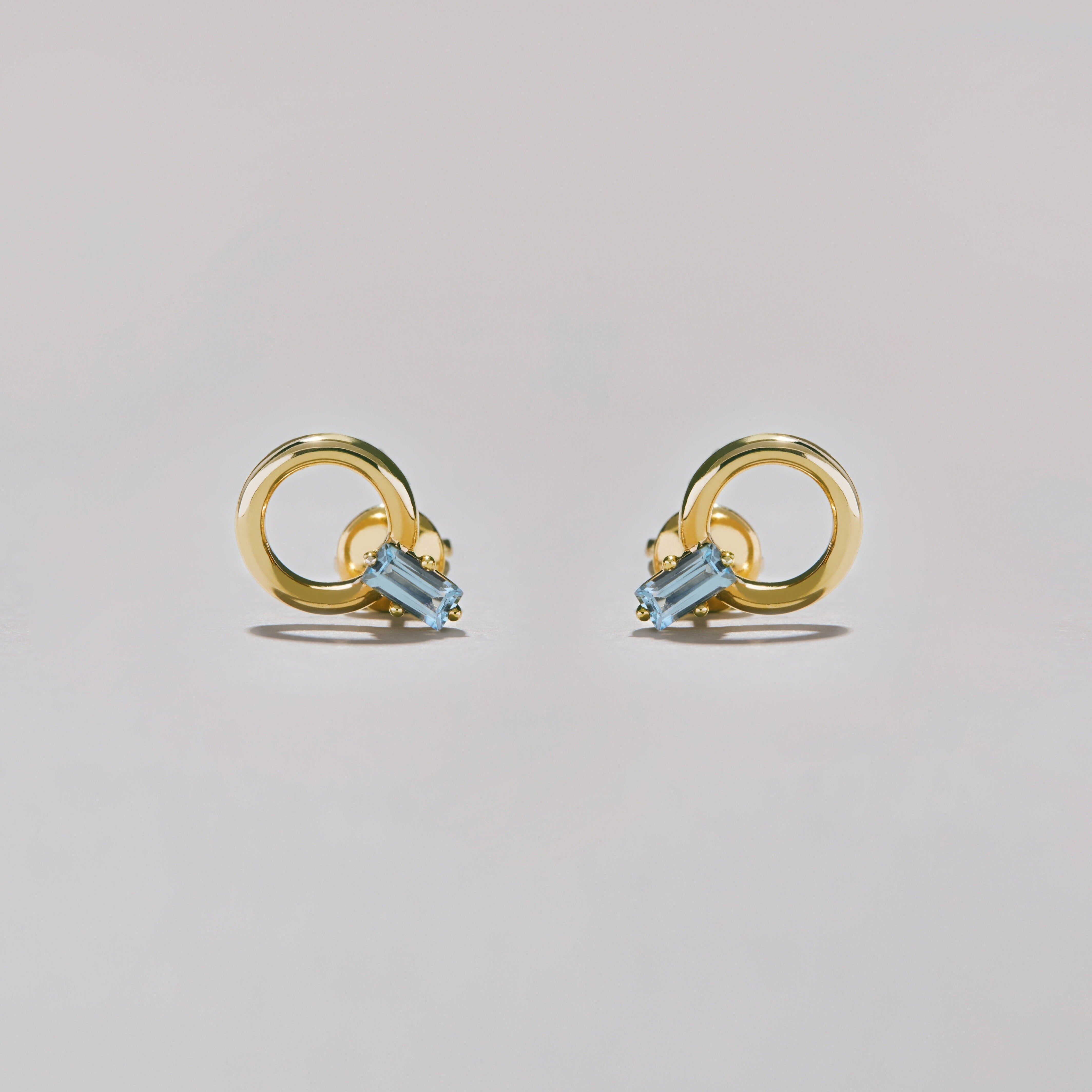 Pair of gold hoop stud earrings with rectangular blue gemstones on soft grey surface.