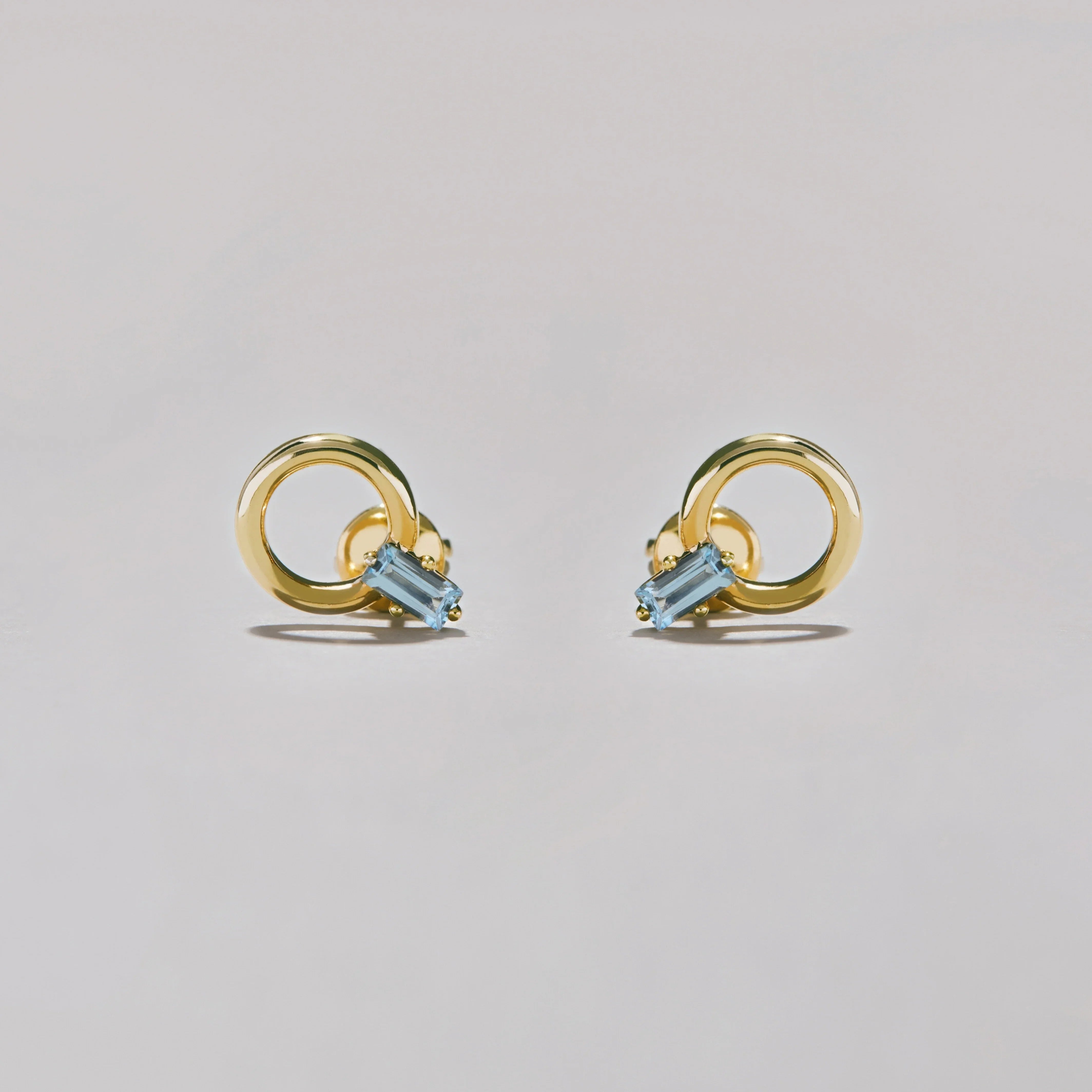 Pair of gold hoop stud earrings with rectangular blue gemstones on soft grey surface.