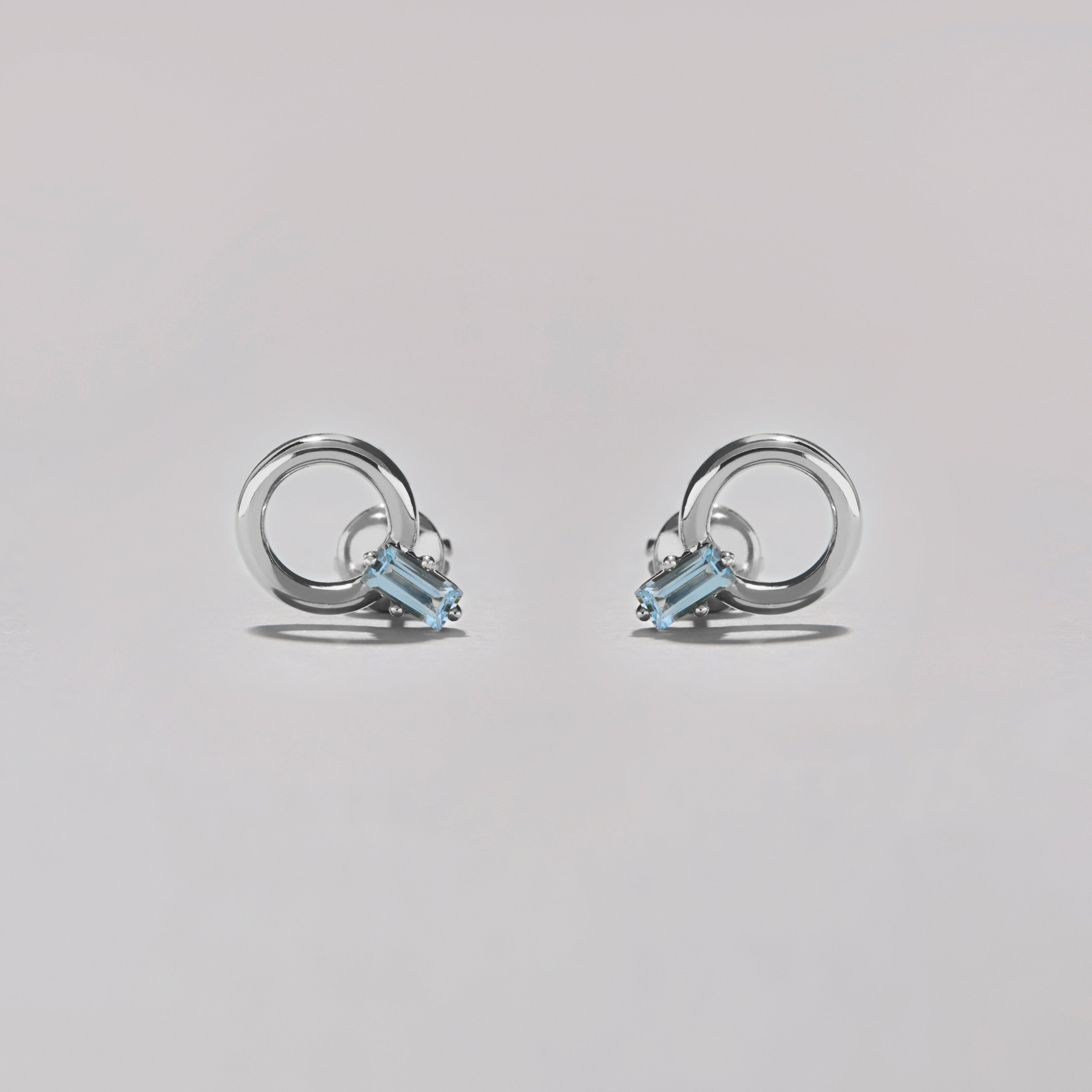 Pair of silver hoop stud earrings with rectangular blue gemstones on soft grey surface.