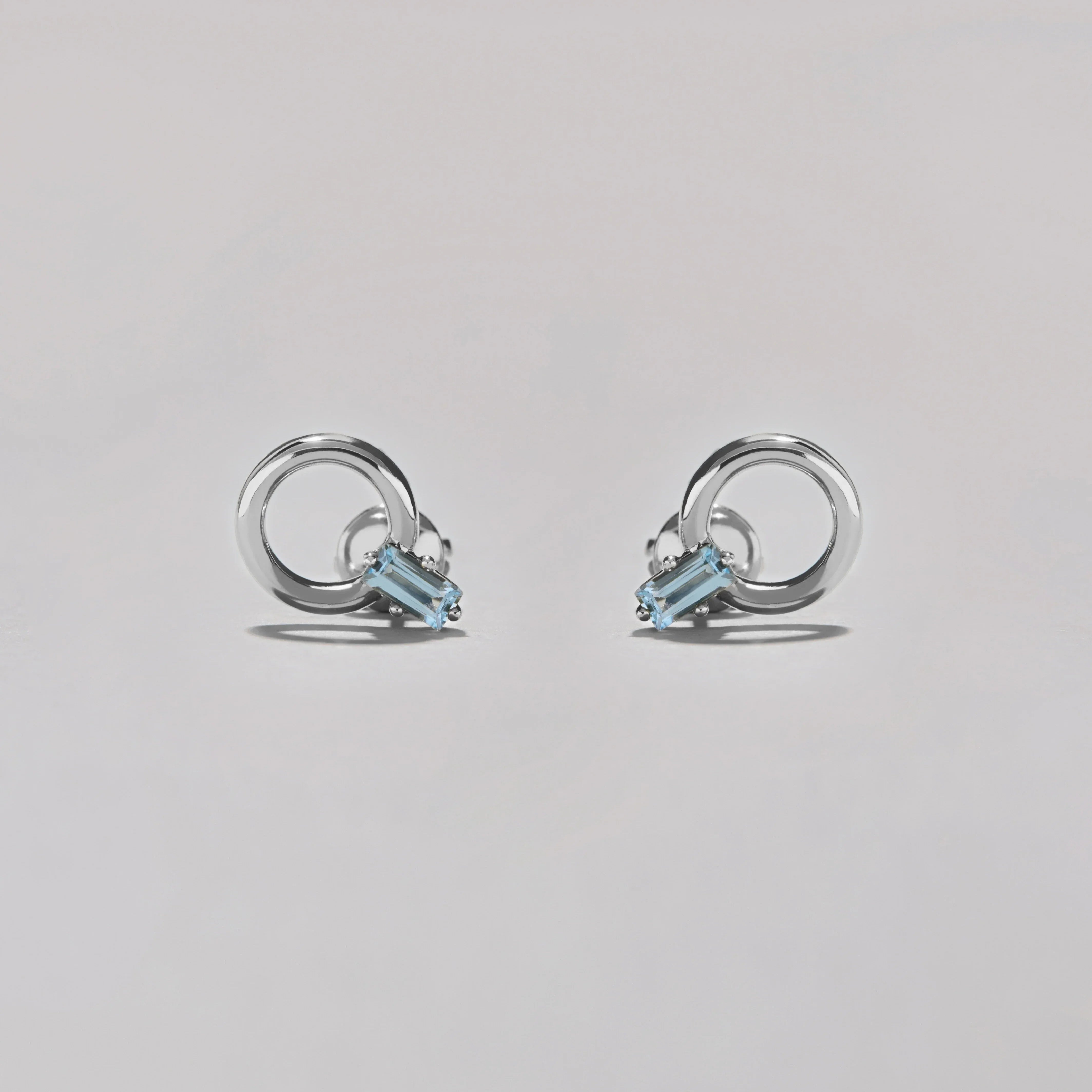 Pair of silver hoop stud earrings with rectangular blue gemstones on soft grey surface.