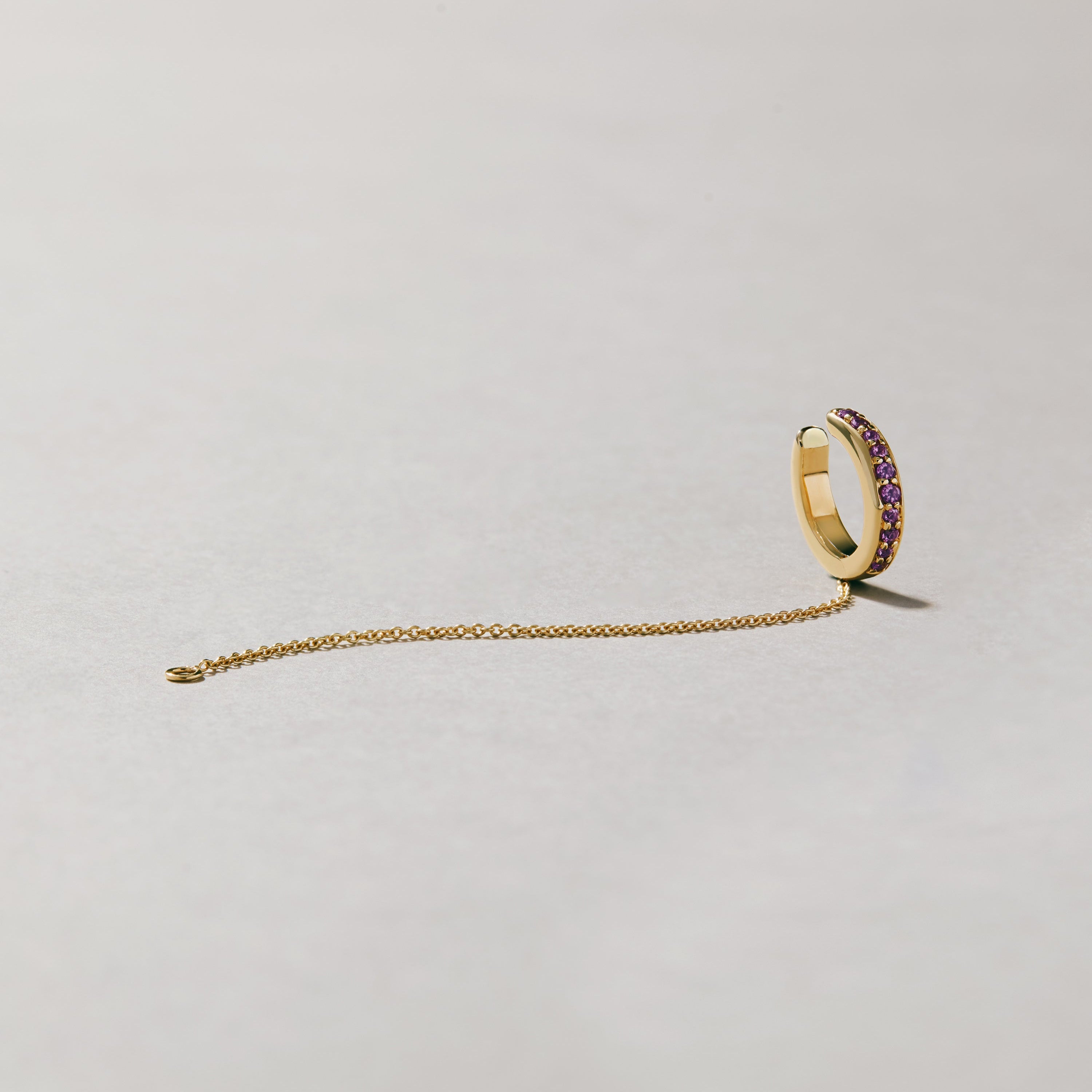 Product image of gold-tone ear cuff with purple gemstones and slim chain laid out on pale background.
