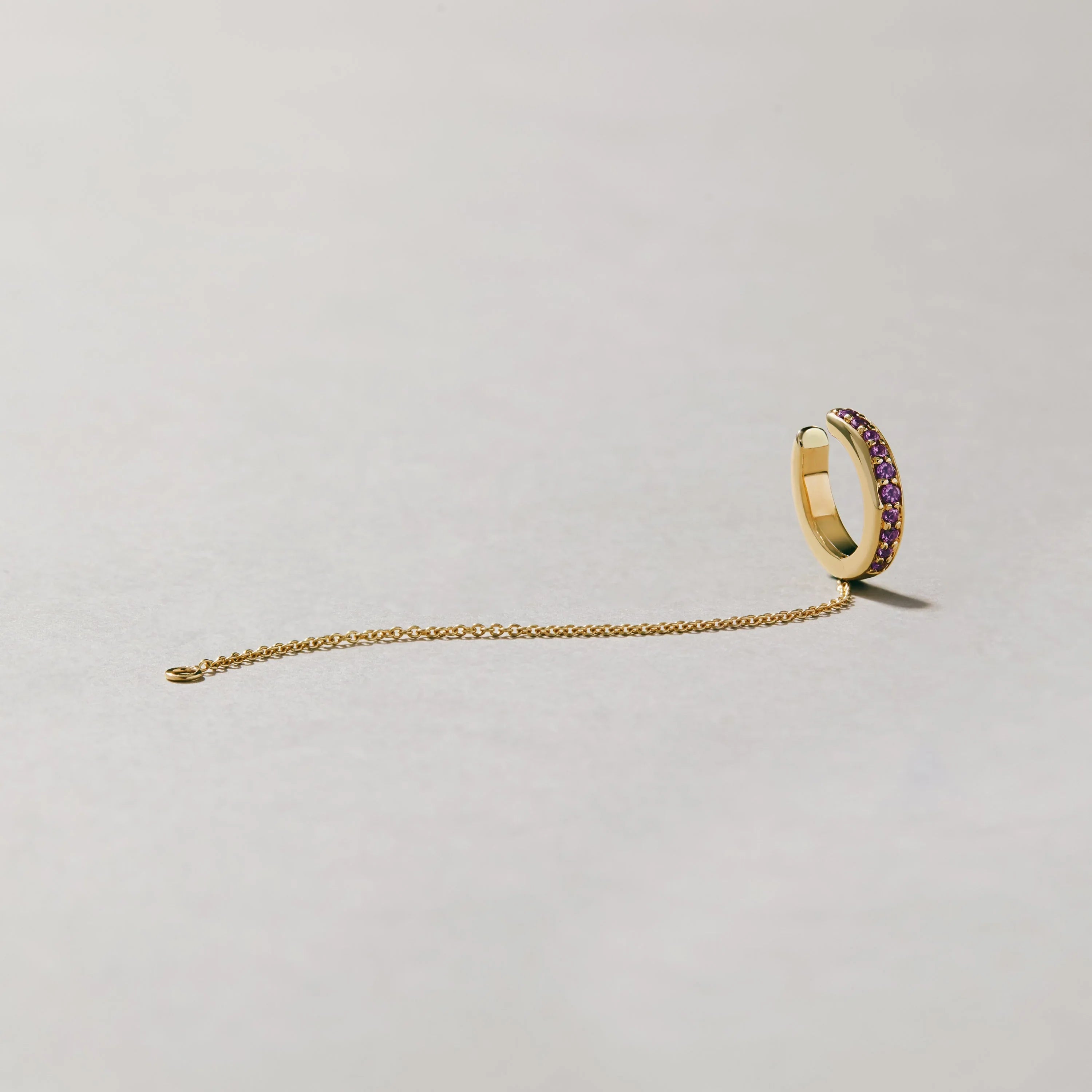 Product image of gold-tone ear cuff with purple gemstones and slim chain laid out on pale background.