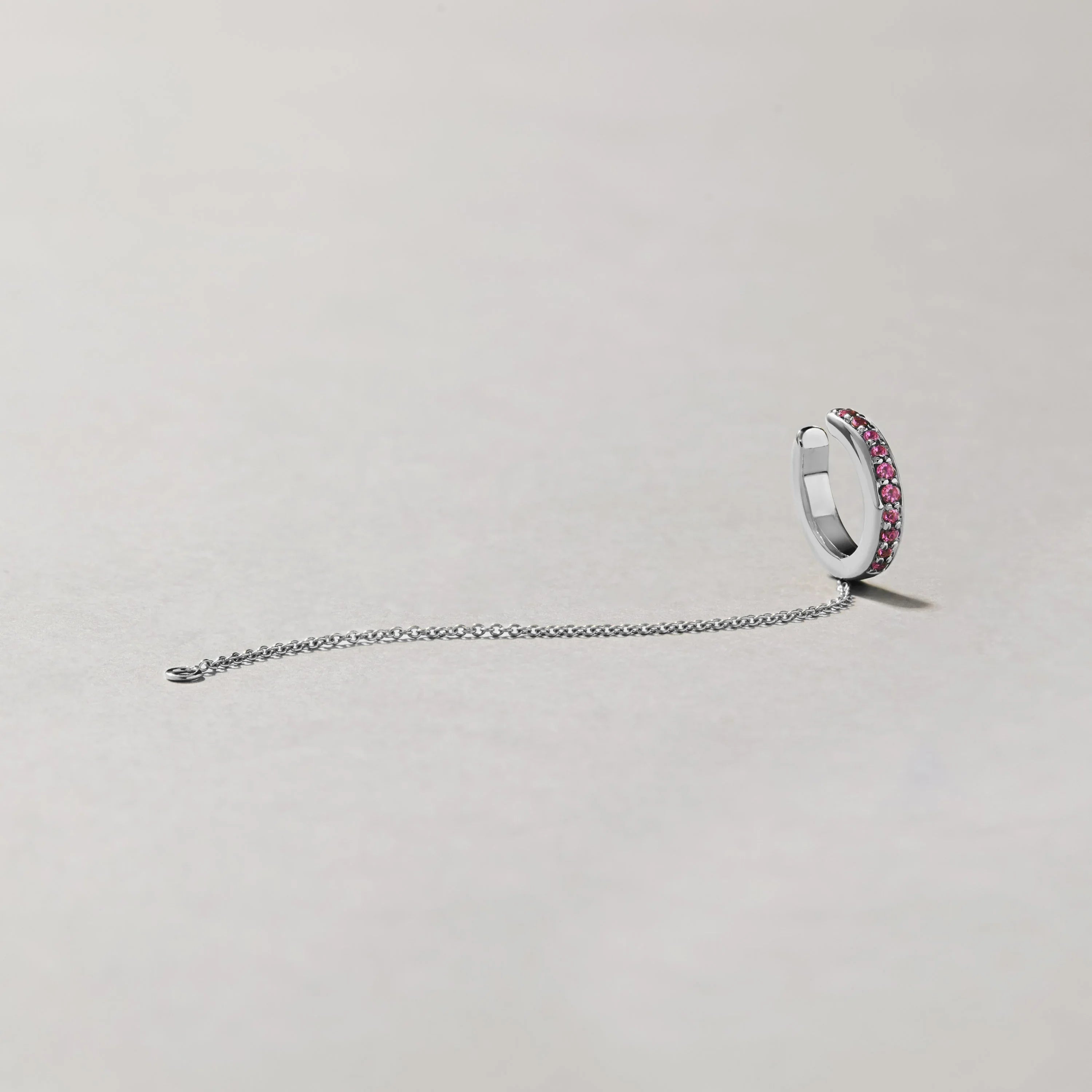 Product image of silver-tone ear cuff with pink gemstones and slim chain laid out on pale background.