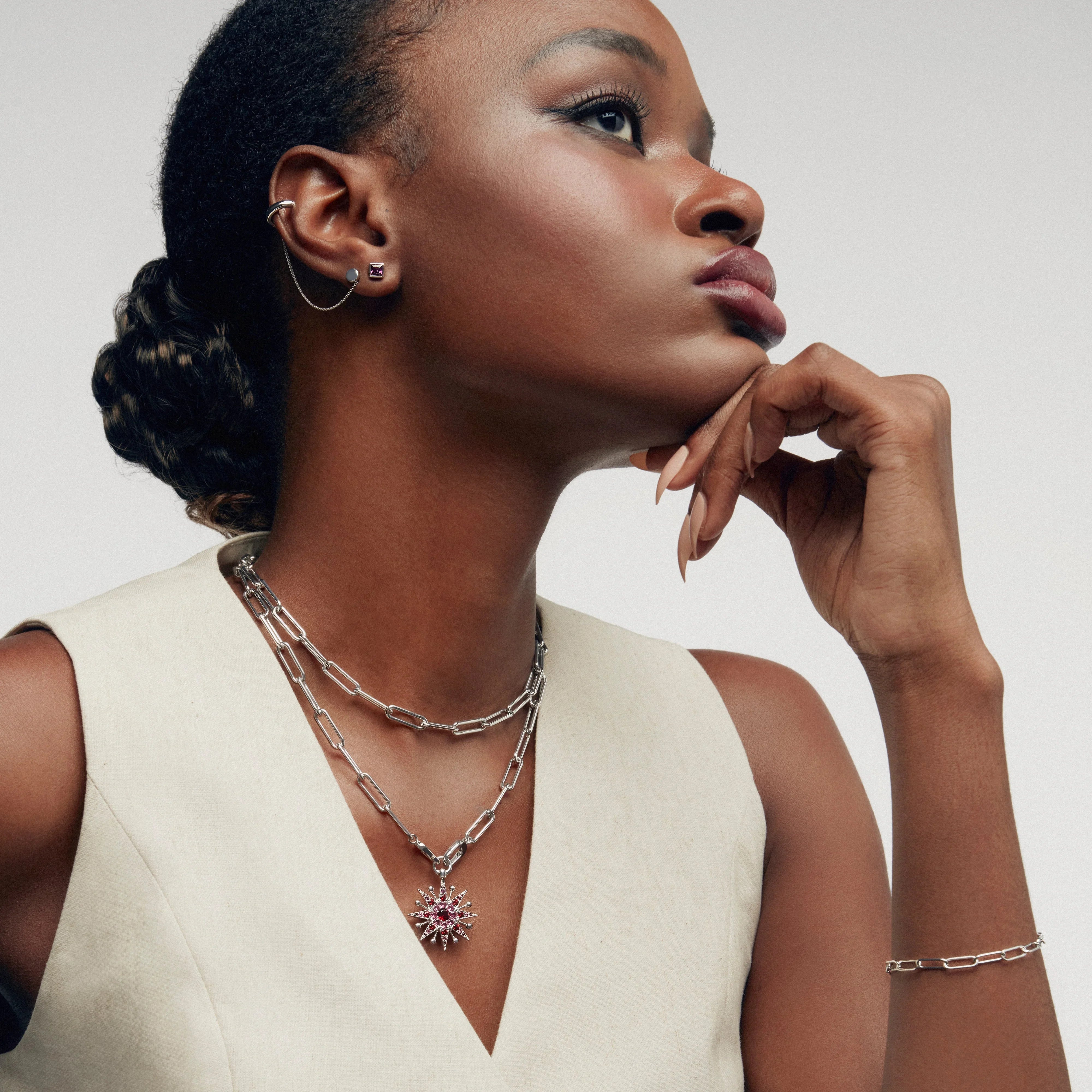 Profile view of a woman with short hair styled in a bun, wearing a sleeveless cream top. She is accessorized with a layered silver chain necklace featuring a pink and red gemstone starburst pendant, a minimalist bracelet, and double earrings connected with a chain.