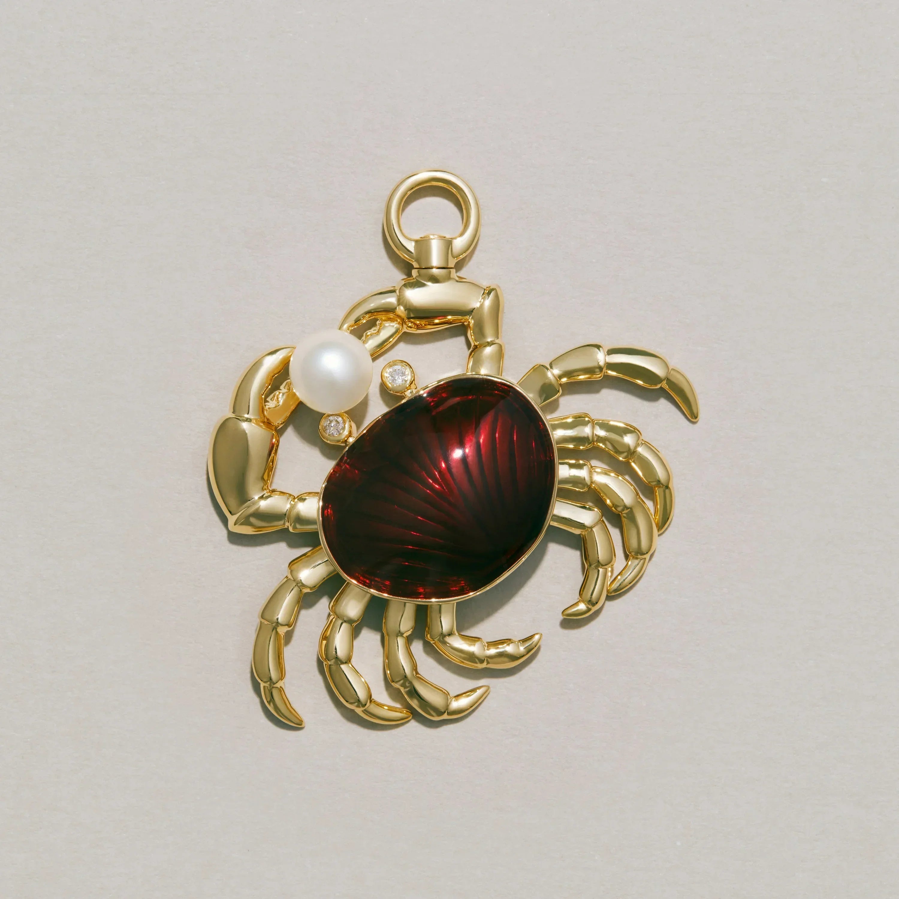 Gold crab-shaped charm with a red enamel and pearl on a light gray background