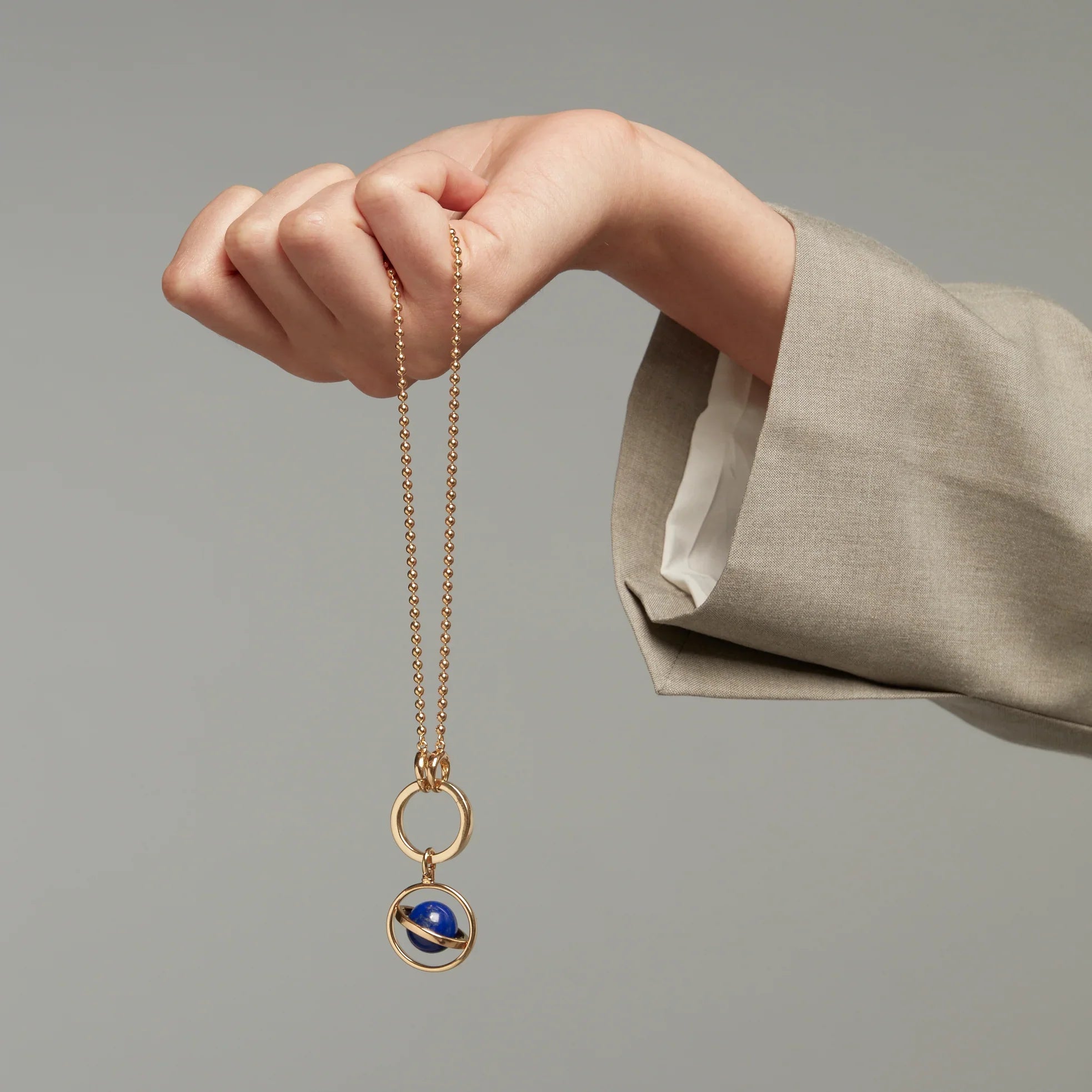 Hand holding a gold chain necklace with a blue planet-like orb pendant against a neutral background.