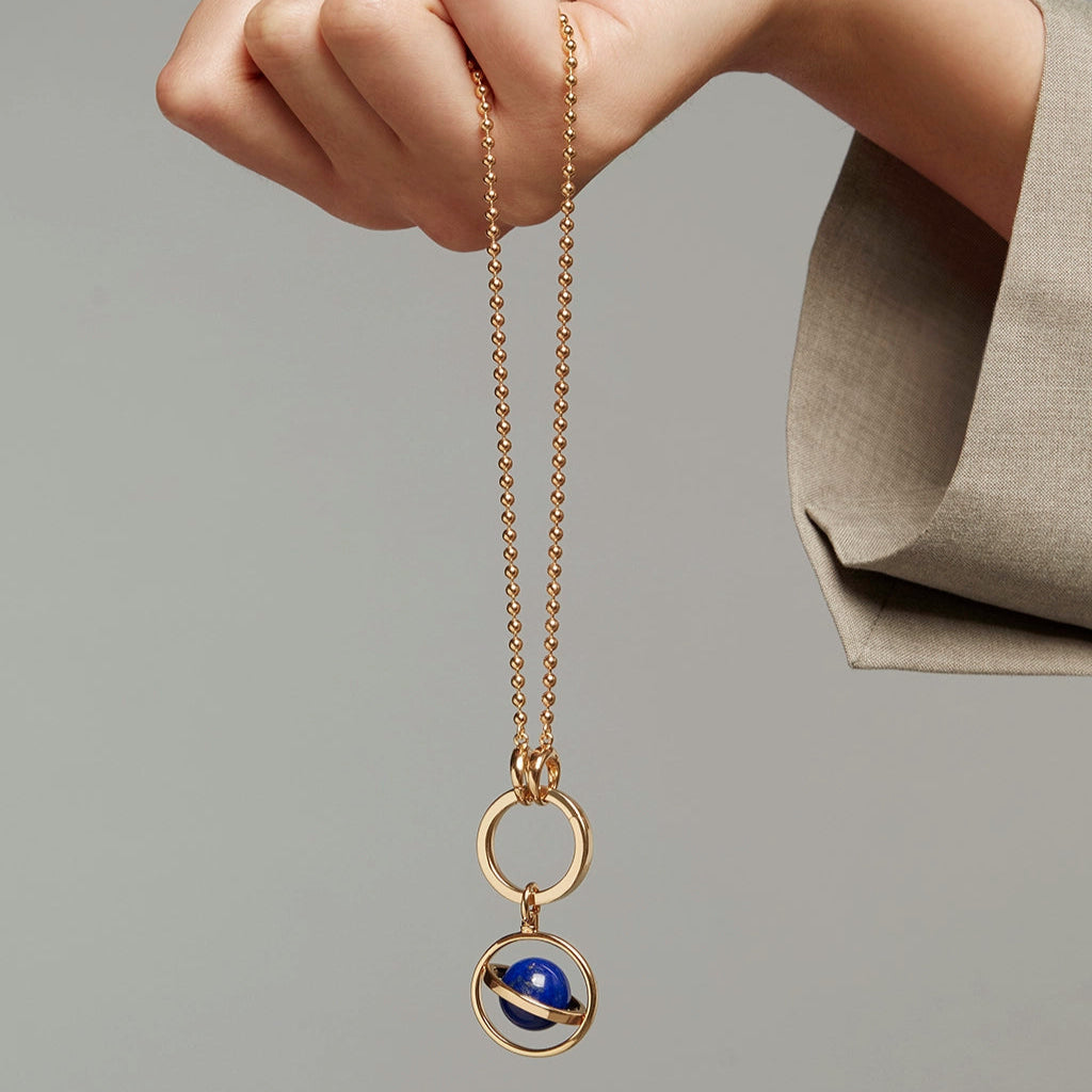 Hand holding a gold chain necklace featuring a rotating planetary charm with a blue gemstone centerpiece.