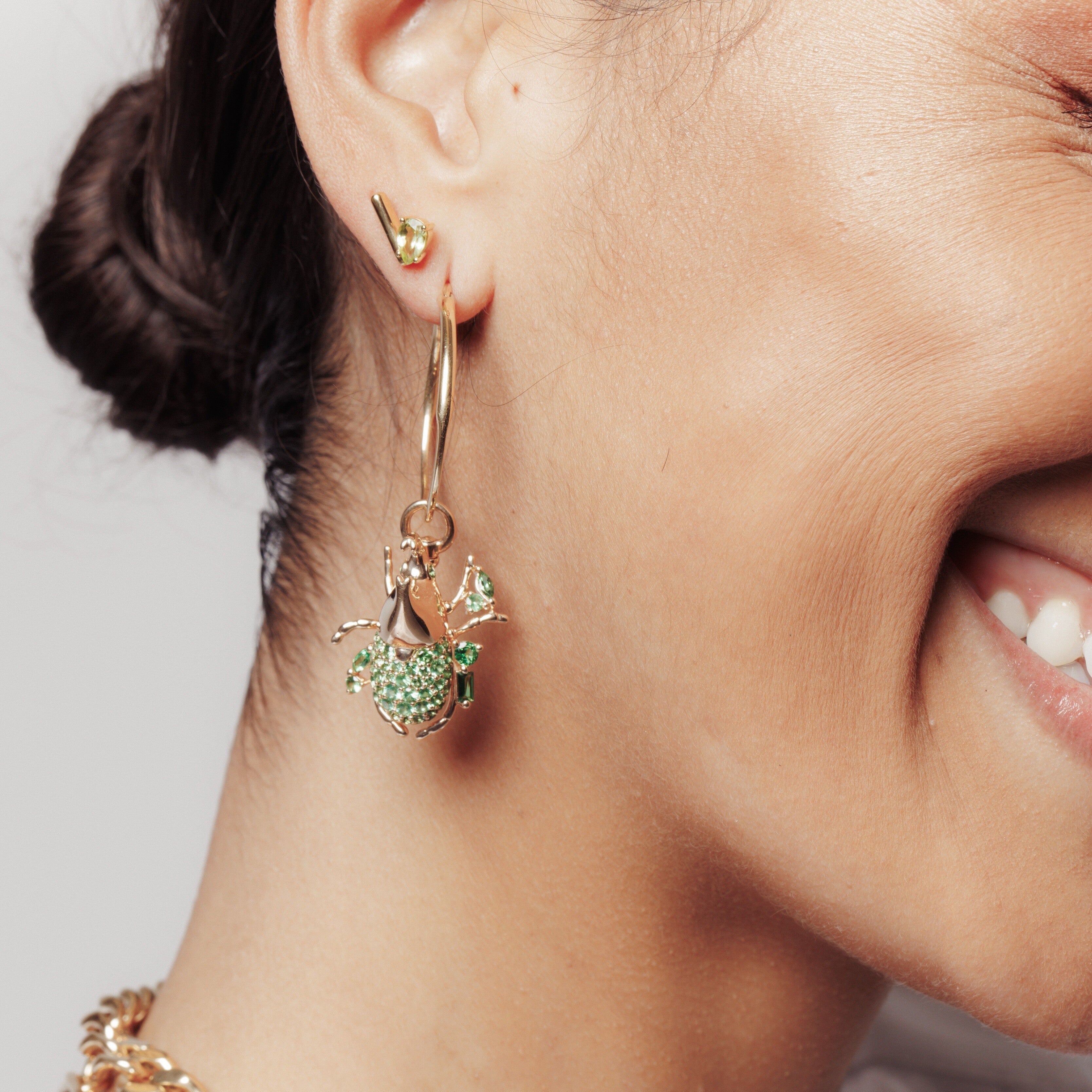 Gold hoop earring with detailed green crystal beetle charm, worn on model's ear.