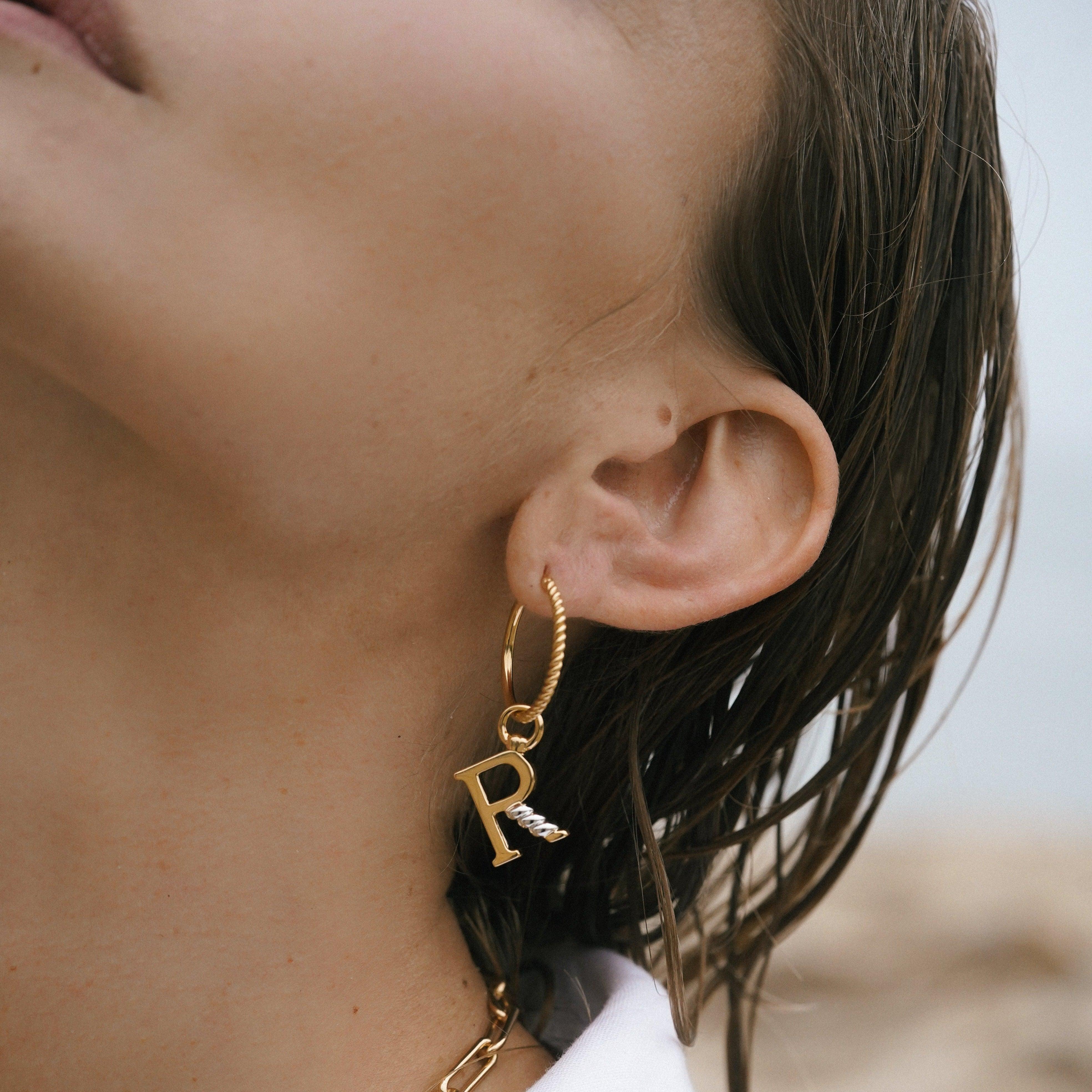 Close-up of gold hoop earring with hanging gold letter R charm featuring a silver rope twist detail.