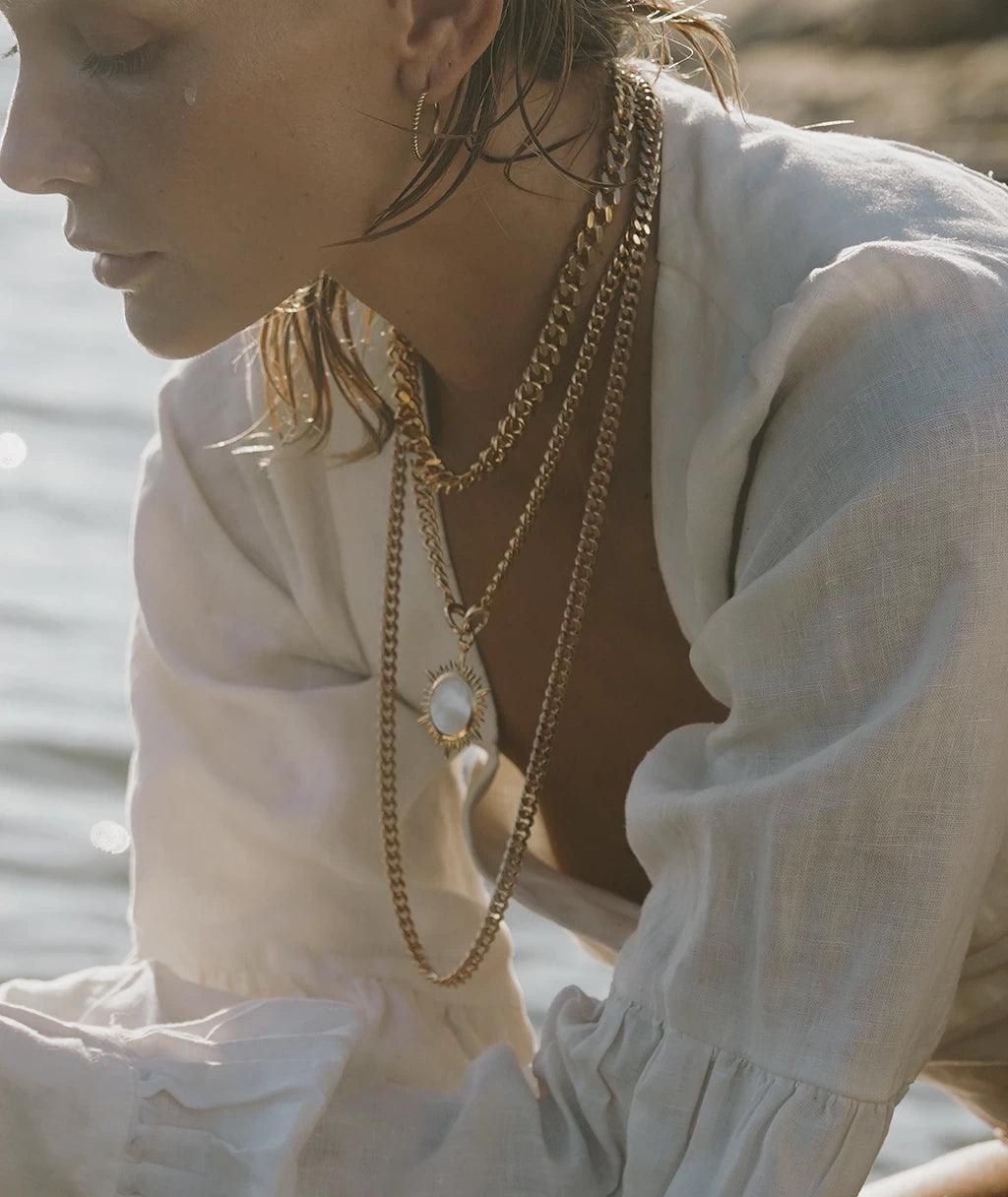 Layered gold charm necklaces styled on model by the sea with white blouse