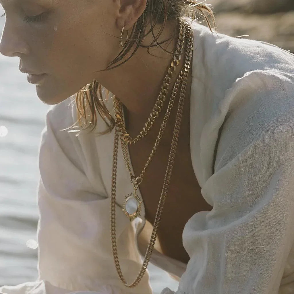 Layered gold charm necklaces styled on model by the sea with white blouse