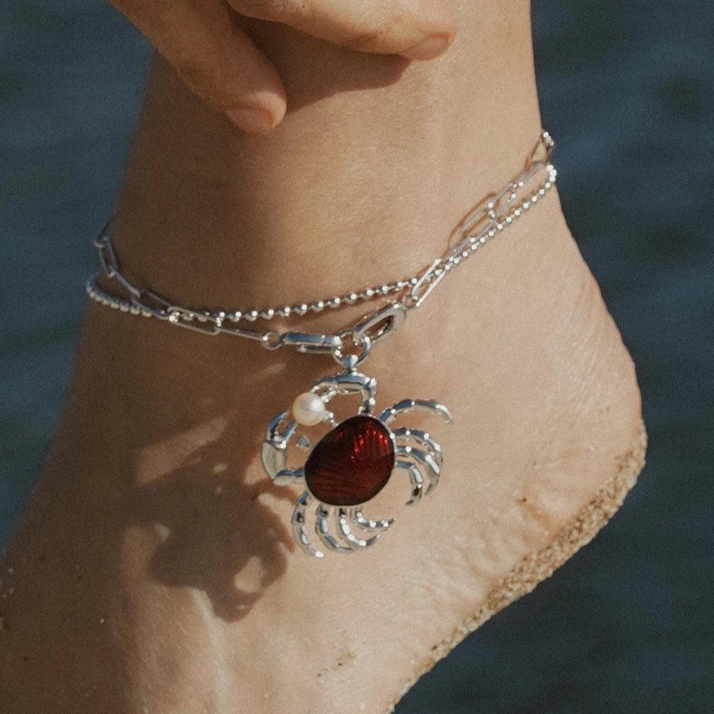 Silver anklet with red enamel crab charm and pearl accent, styled on a sandy beach.