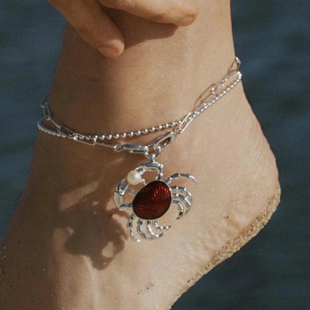 Silver anklet with red enamel crab charm and pearl accent, styled on a sandy beach.