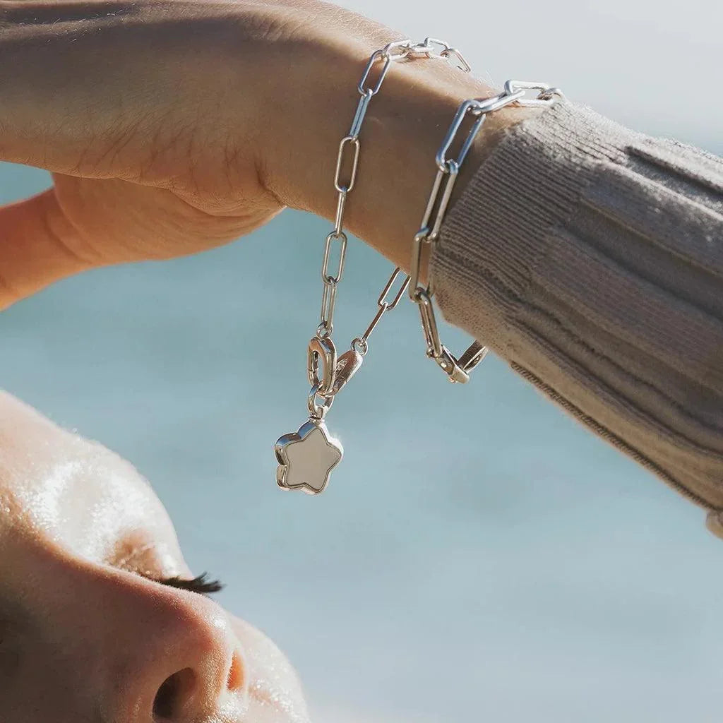 Silver paperclip bracelet with star charm draped on wrist against a coastal backdrop.