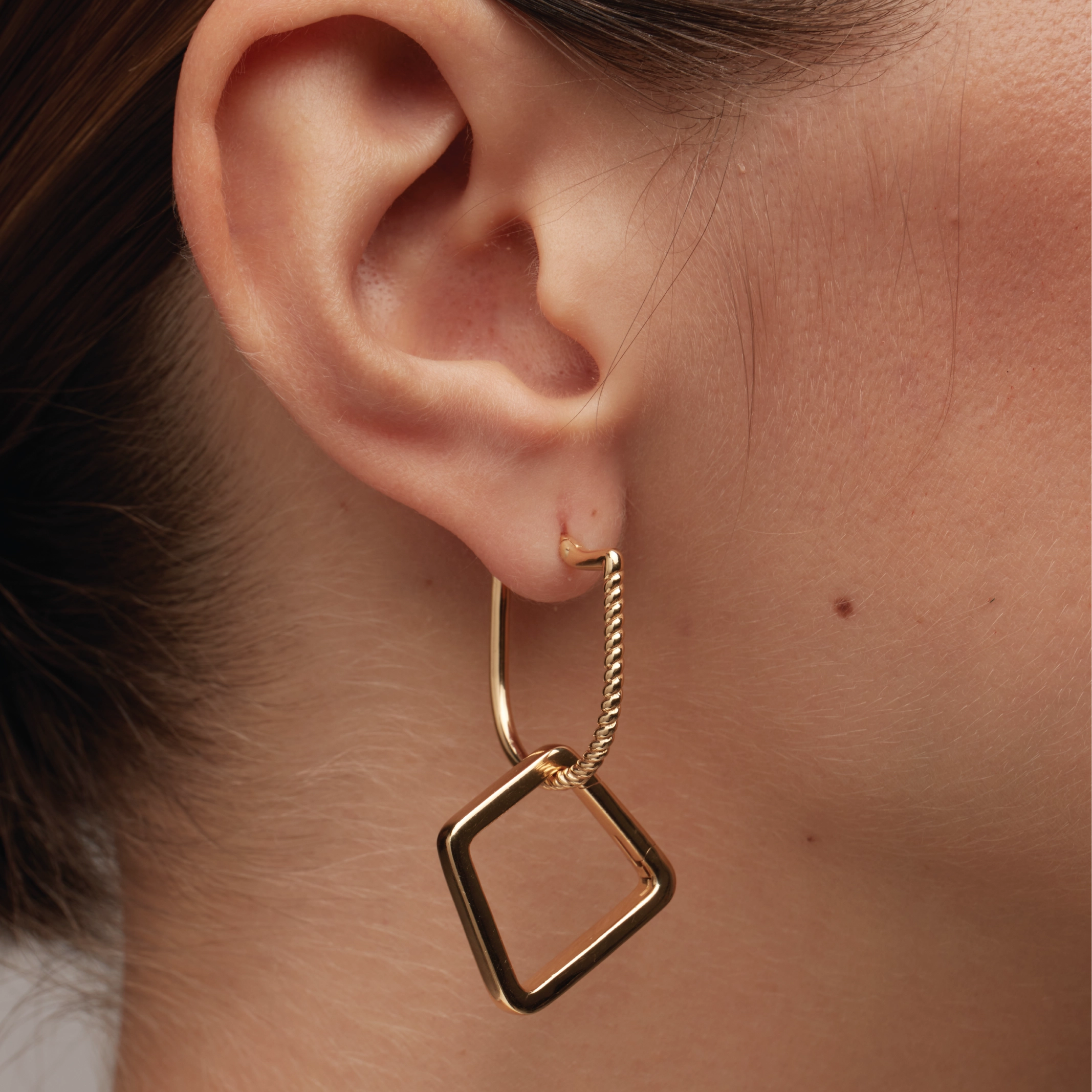 Close-up of an ear wearing a gold hoop earring with a twisted rope detail and a minimalist hollow diamond-shaped gold charm hanging below.