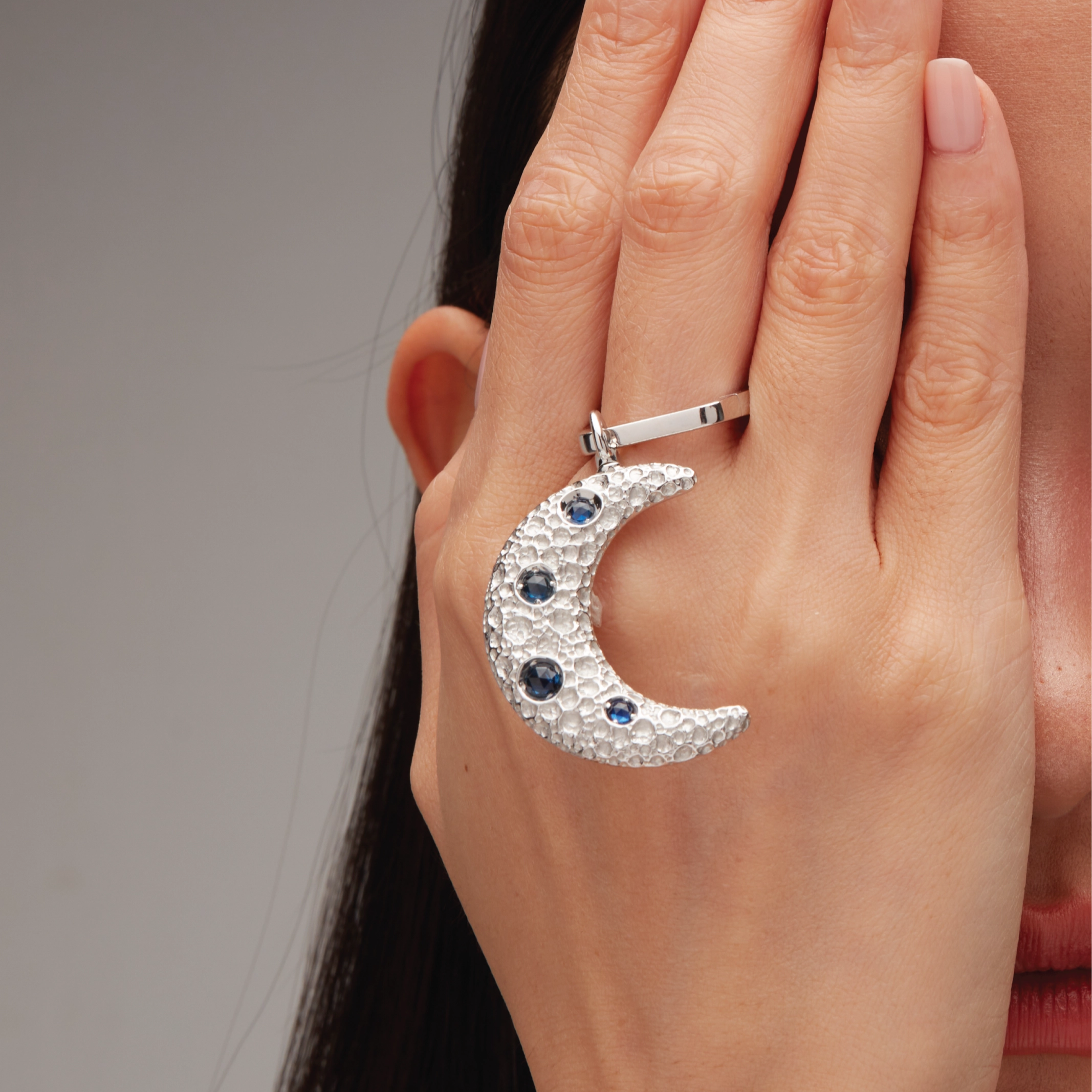 Silver crescent moon charm with blue gemstones, hooked onto a sleek silver ring, held against the side of the face.