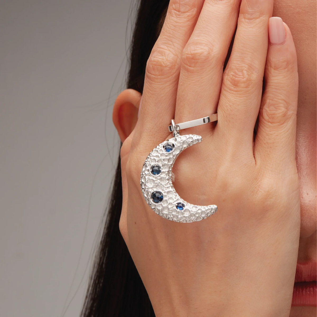 Silver crescent moon charm with blue gemstones, hooked onto a sleek silver ring, held against the side of the face.