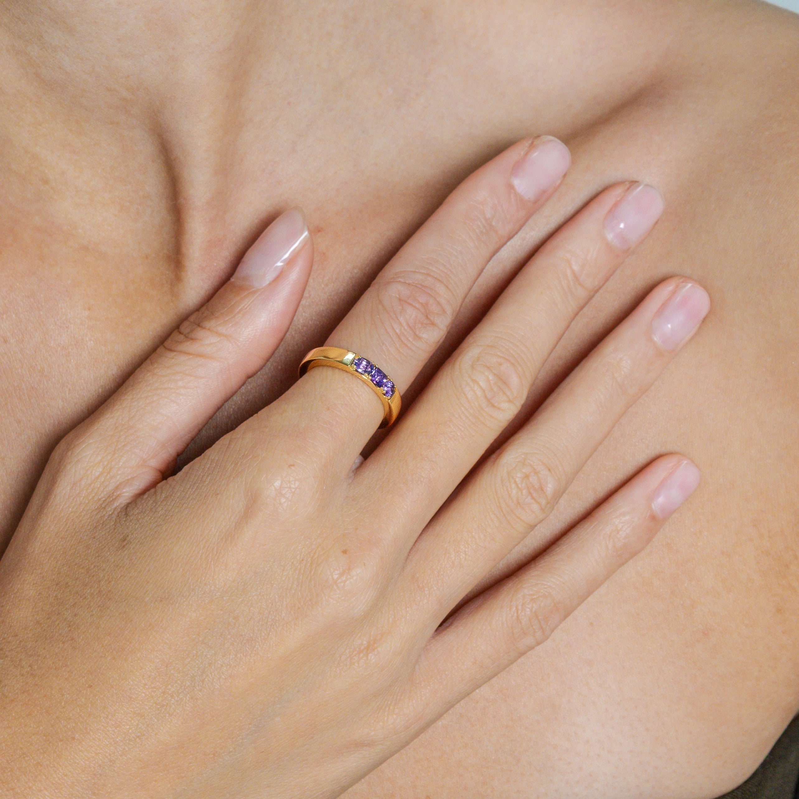 Close-up of a hand wearing a gold ring with a purple gemstone on a neutral background