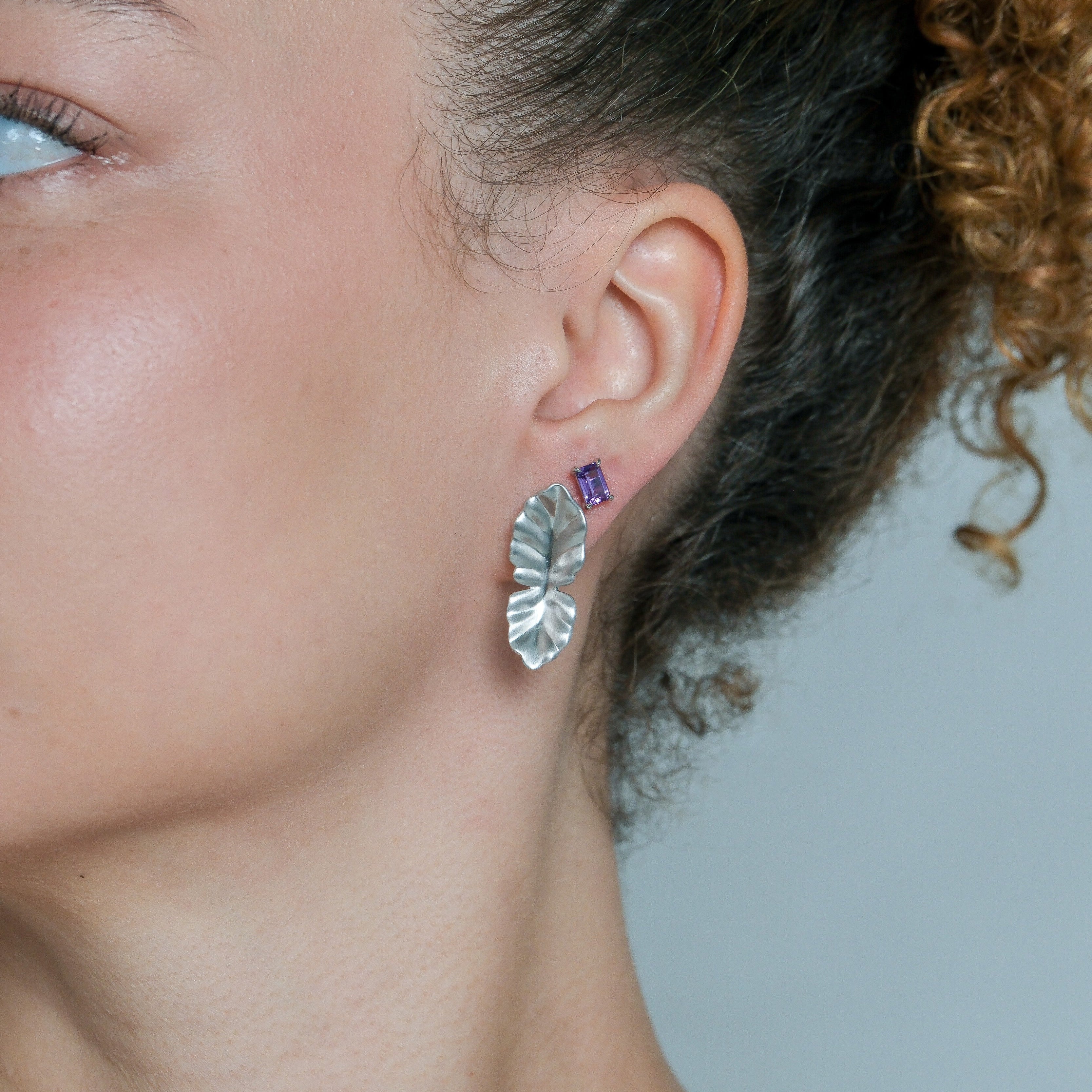 Close-up of a person wearing a silver leaf-shaped earring and a purple gemstone stud against a neutral background