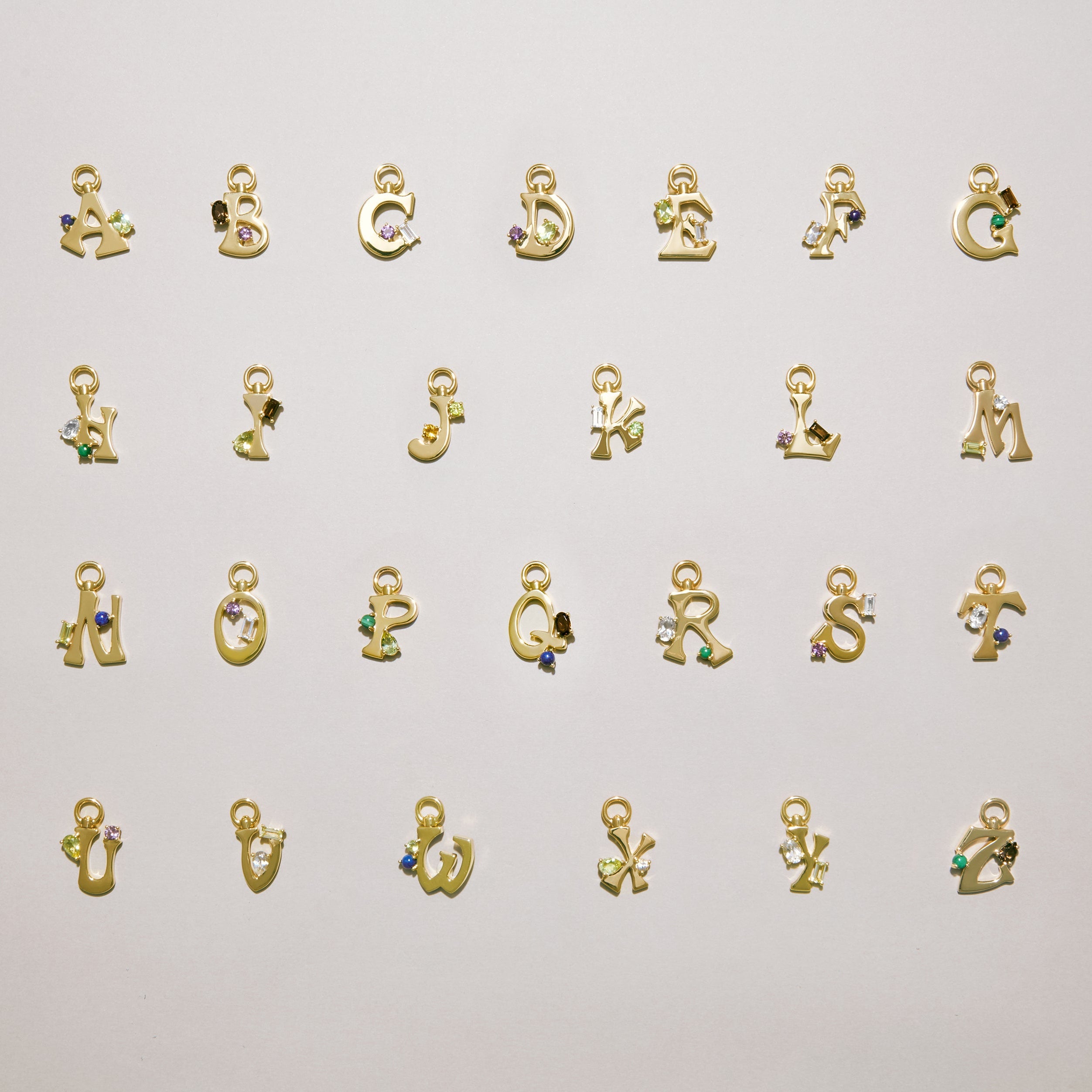Alphabet of gold letter charms A–Z laid out on a light background, each set with colorful gemstones.